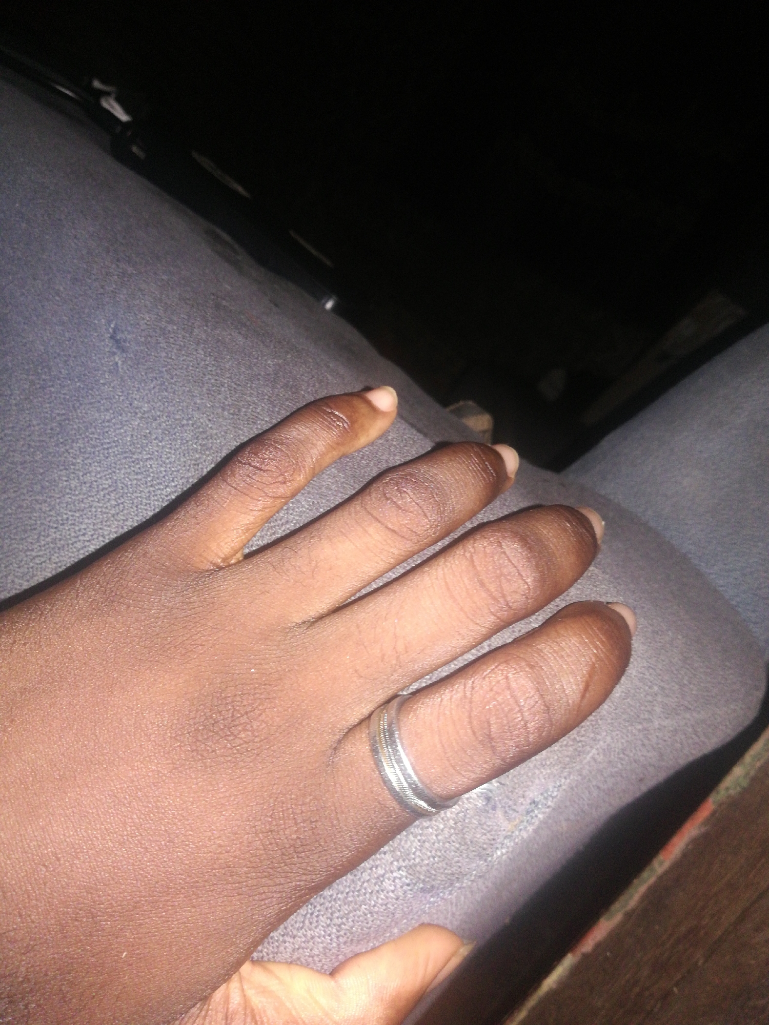 A close-up shot of a dark-skinned hand with a silver ring on the ring finger. The hand rests on a blue fabric surface. The background is dark and out of focus. The image provides no information about the location in Lagos, Nigeria.