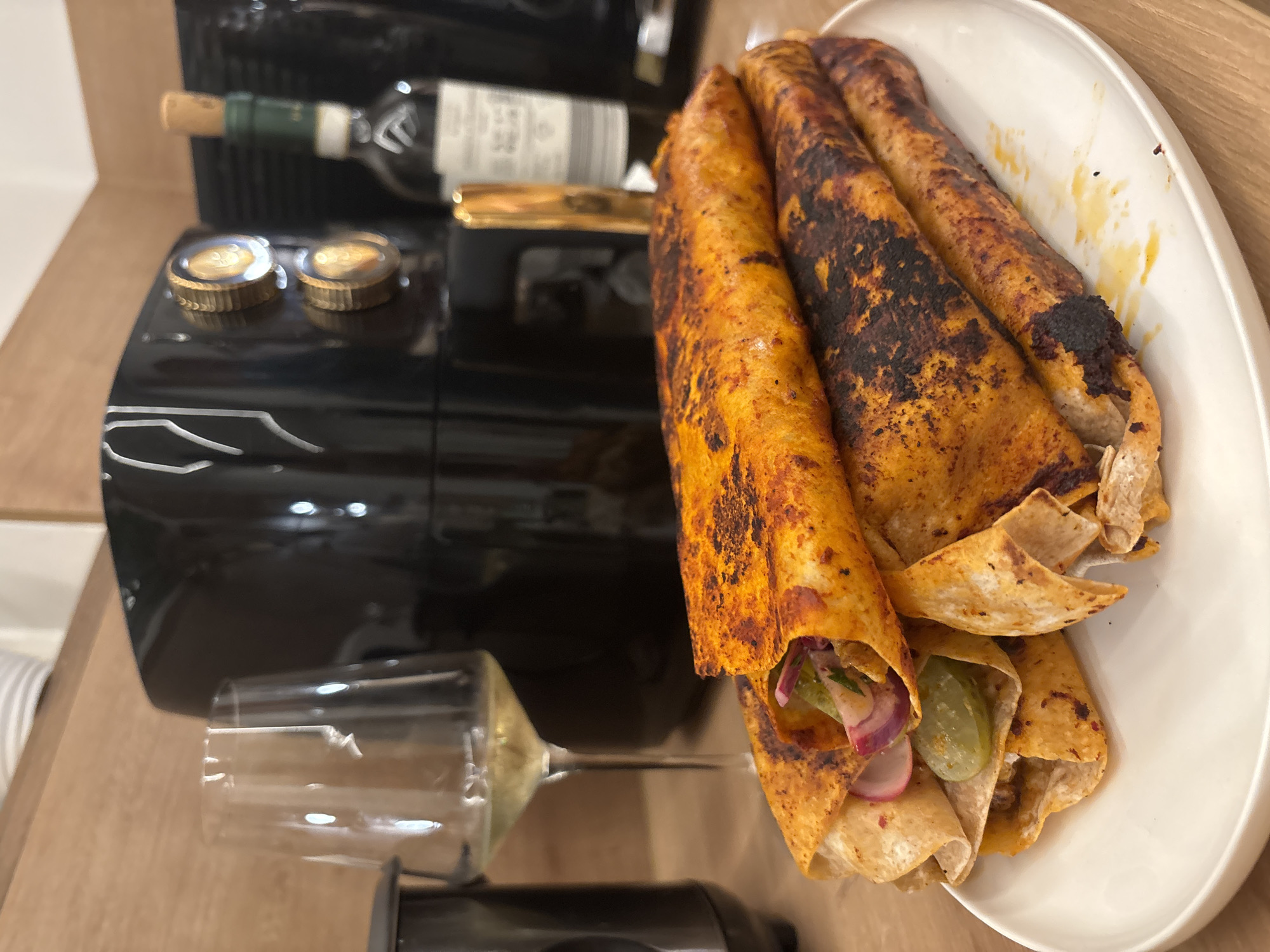 The image depicts a plate of taquitos, possibly al pastor given the reddish-orange hue of the tortillas, stacked on a white plate. The taquitos appear to be filled with some sort of meat and garnished with thinly sliced red onion, radish, and possibly pickles. The tortillas are slightly charred in places, suggesting they have been fried or cooked at high heat. In the background, an air fryer is visible, along with a wine glass partially filled with white wine and a bottle of wine with a cork. The setting appears to be a kitchen counter made of wood. There is no indication of the location in Viernheim, Germany.