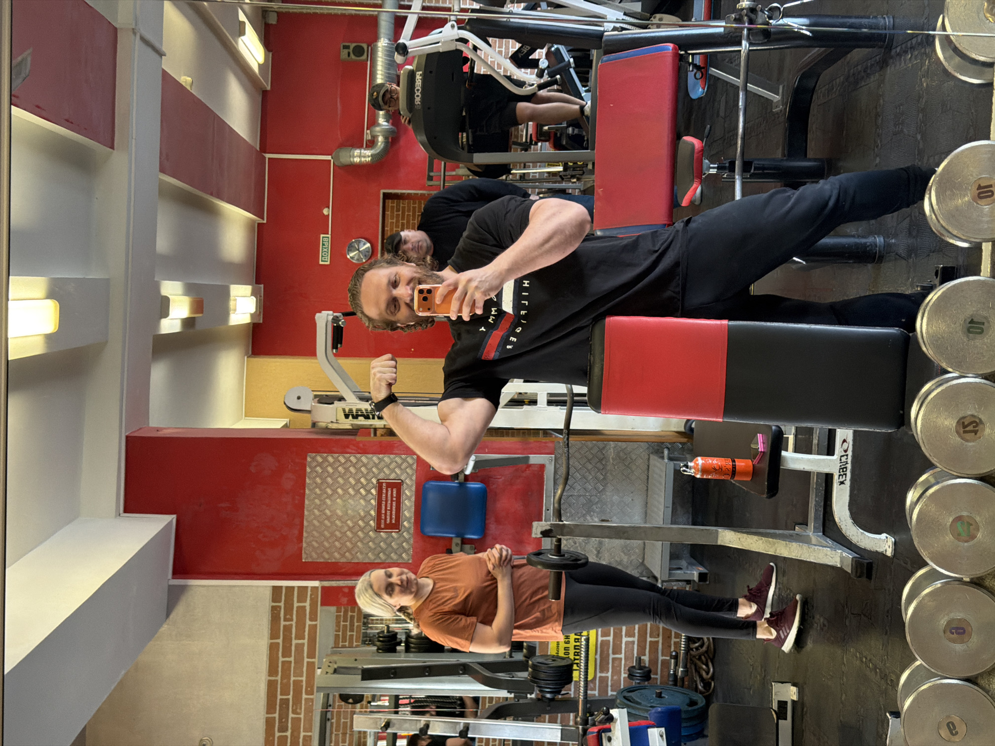 The image depicts a man taking a selfie in a gym. The man is lying on a weight bench with his legs extending towards a rack of weights. He is flexing his bicep and holding an orange cell phone. The reflection in the mirror shows the man and other gym equipment. A woman stands in the foreground and is looking to the side with her hands clasped in front of her. Gym equipment and weights are visible throughout the gym. The gym has red and white walls, with some brick accents. The writing on the wall is Cyrillic, which helps to identify the location as Saint Petersburg, Russia.