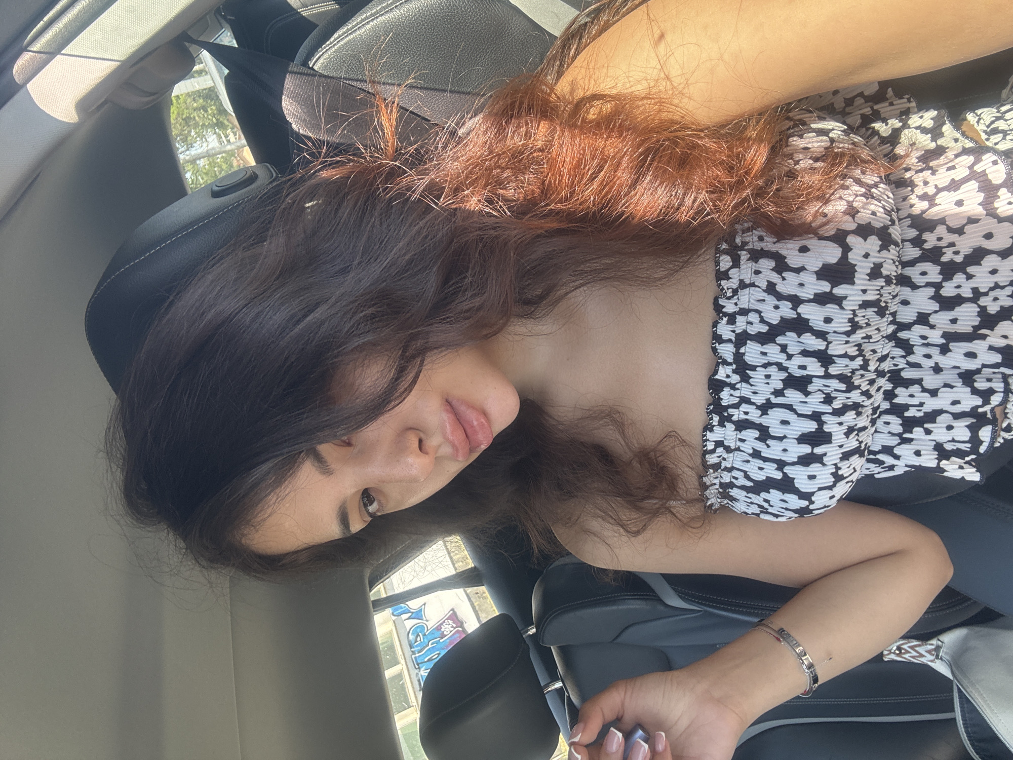 A young woman with long, dark brown and auburn hair is lying down in a car, viewed from a slightly elevated angle. She is wearing a black and white floral print top. Her right arm is extended, and she is wearing bracelets on her wrist. Her left arm is bent, with her hand near her mouth. The sun is shining brightly, casting strong shadows and highlighting her hair. The background shows car seats, a window with trees and buildings visible outside, and a portion of a graffiti-covered wall that reads "ART." The overall mood appears casual and relaxed. The time of day is likely daytime due to the bright sunlight. The location is Plovdiv, Bulgaria, as indicated by the graffiti which includes Cyrillic script.