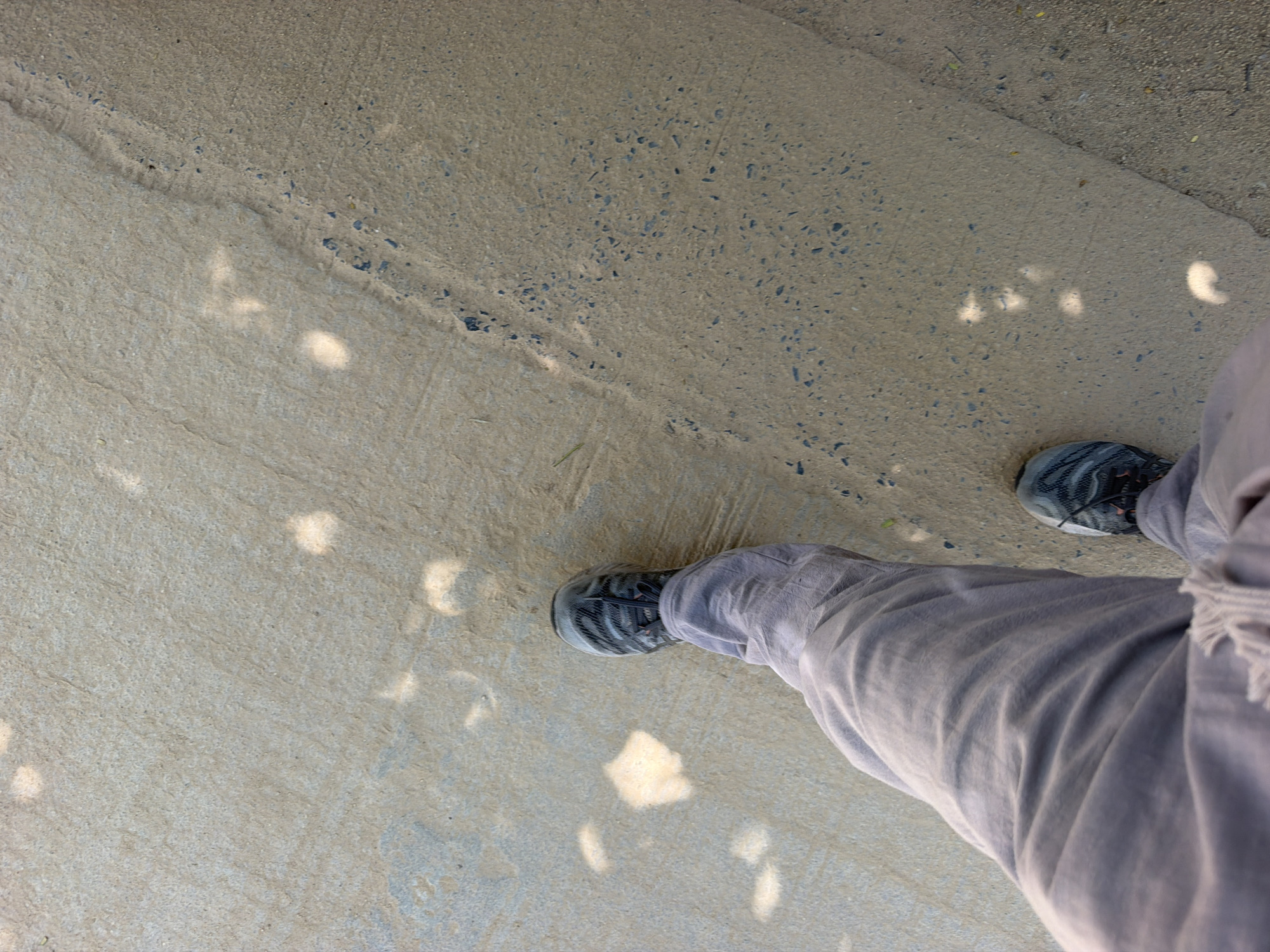 A first-person perspective shot shows a person walking on a light-colored, textured surface that appears to be pavement or concrete. The surface is uneven and has small, dark pebbles embedded in it. Sunlight casts dappled shadows on the ground, creating irregular shapes of light and dark. The person's left leg, clad in light grey, loose-fitting pants, is visible in the lower left of the frame. A grey and black athletic shoe is on their left foot. The right shoe and leg are also partially visible, indicating the person is walking forward and slightly to the right. The overall environment suggests an outdoor urban or semi-urban setting.