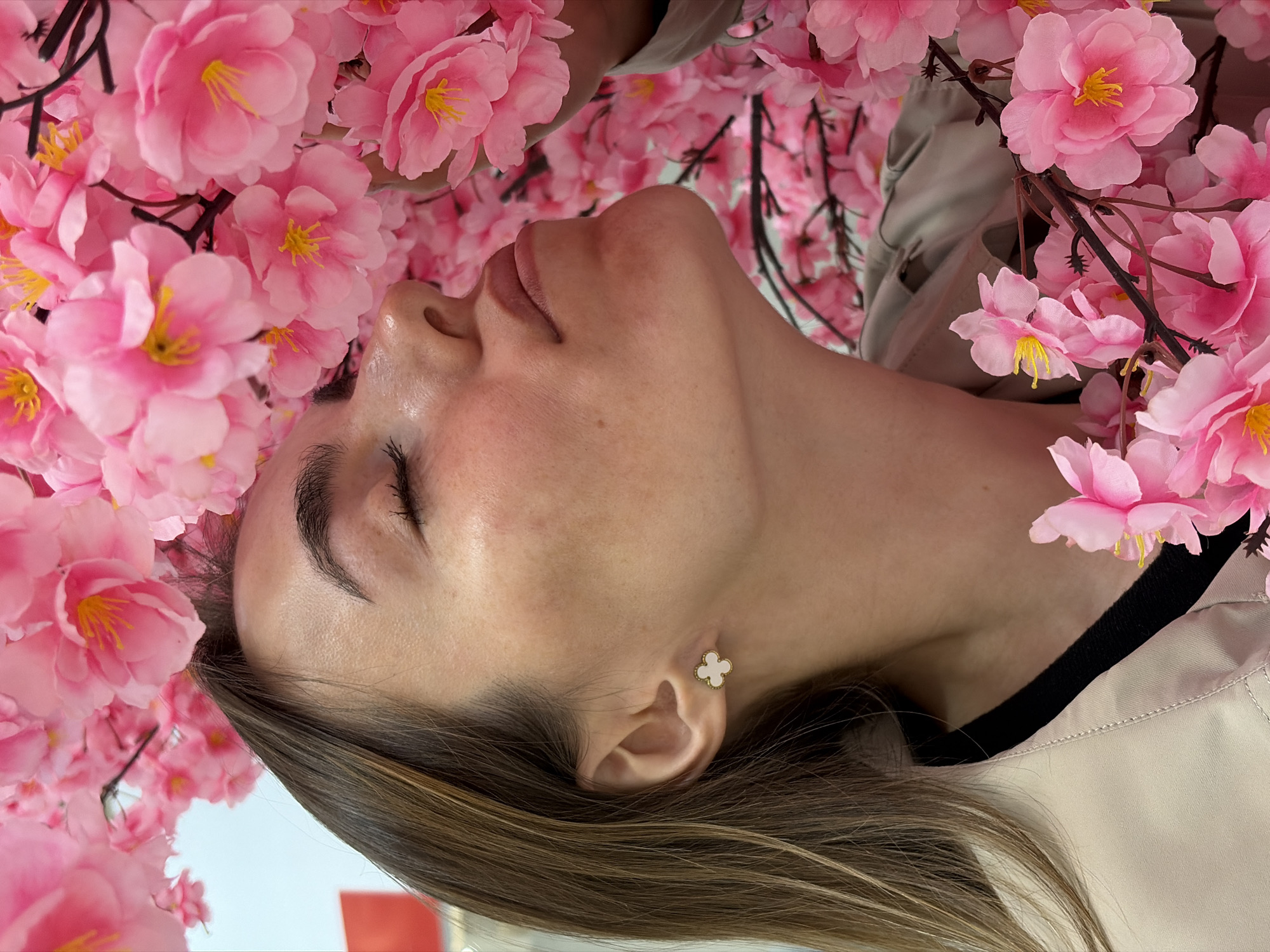 A woman lies among pink flowers, possibly cherry blossoms, with her head tilted back. She has fair skin, dark eyebrows, and closed eyes. She wears a small flower-shaped earring. Her hair is dark brown with lighter streaks, and she is wearing a beige or light-colored top over a dark garment. The background is filled with pink flowers, creating a lush and immersive setting. The image was taken upside down. There is no indication this image was taken in Izhevsk, Russia.