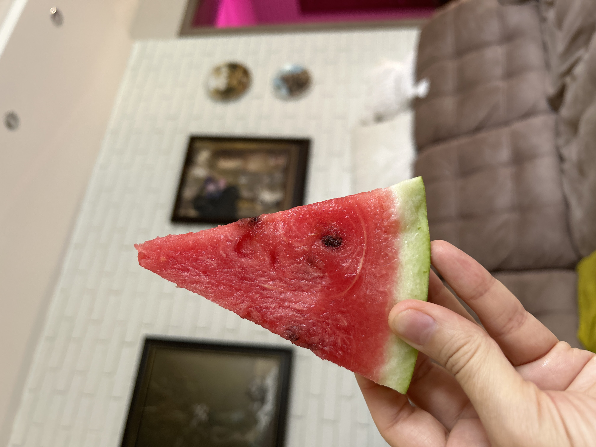 A close-up, shallow depth-of-field shot shows a hand holding a triangular slice of watermelon. The watermelon is bright red with a thin green rind, and it appears to be freshly cut. The hand is pale with visible veins and fingernails, and the skin texture is slightly wrinkled. In the background, a white brick wall is visible, with two framed pictures hanging on it. The picture on the left is darker and appears to be a landscape or portrait, while the one on the right is smaller and features circular elements. Further in the background, a brown tufted sofa is partially visible, and to the right, a bright pink light source casts a glow. The scene appears to be indoors, likely in a residential setting, and there is no indication of the city of Минино, Russia.