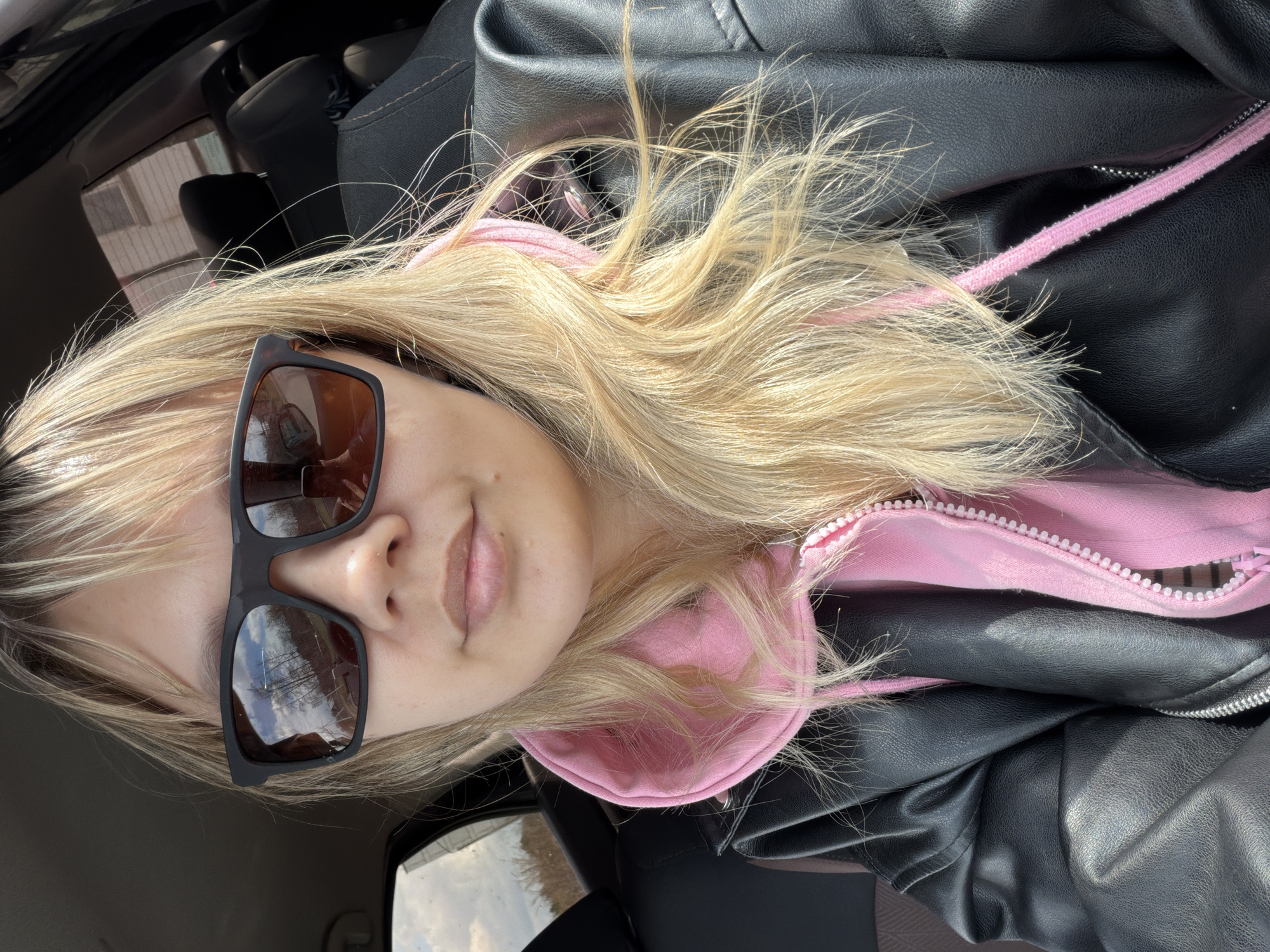 The media file displays a close-up portrait of a person, likely female, with long, wavy blonde hair. The image is oriented horizontally, with the subject's head positioned on the left side. She is wearing dark sunglasses that cover her eyes, a pink hooded sweatshirt with a white zipper, and a black leather jacket over it. The sunglasses have rectangular frames and reflective lenses that show a reflection of trees and a cloudy or partly cloudy sky, indicating an outdoor setting.
The subject's face is partially visible, with her nose and mouth area showing a neutral or slightly relaxed expression. Her blonde hair is prominent, appearing bright and somewhat windswept, framing her face and cascading over her shoulders. The pink of the hoodie provides a contrast to the black leather jacket, which has a textured, slightly wrinkled appearance. A pink drawstring from the hoodie is also visible.
The background suggests the interior of a vehicle, possibly a car, with dark-colored seats and parts of the dashboard or door panel visible. The lighting is bright and natural, indicating it is daytime. No visible text is present in the image.