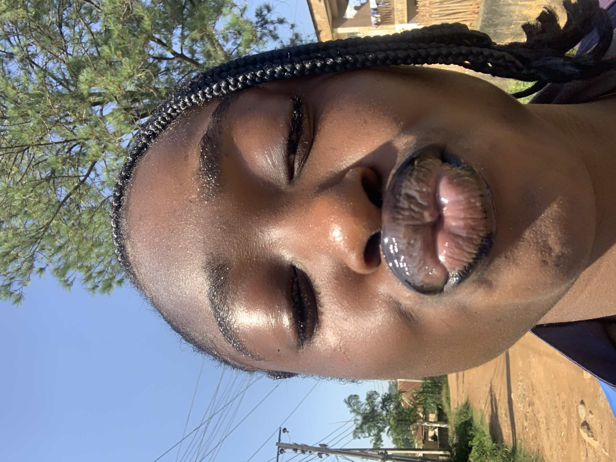 A close-up, slightly angled selfie of a Black female, likely in her late teens or early twenties, is captured outdoors on a sunny day. The subject is puckering her lips, which are coated in a dark, glossy lipstick, reflecting the bright sunlight. Her eyes are closed, and her eyelashes are visible. She has dark, braided hair. Her skin appears dewy and highlighted, possibly from sweat or a luminous cosmetic product.
The background suggests a rural or semi-urban setting. To the left, a dirt road lined with green vegetation leads towards a cluster of buildings, including one with a corrugated metal roof. Utility poles and wires are also visible. To the right, a partially constructed building with a balcony is seen, and beyond that, more trees and buildings. The sky is a clear, bright blue. The overall impression is a candid moment captured in natural light. The location is identified as Kabong, Nigeria.