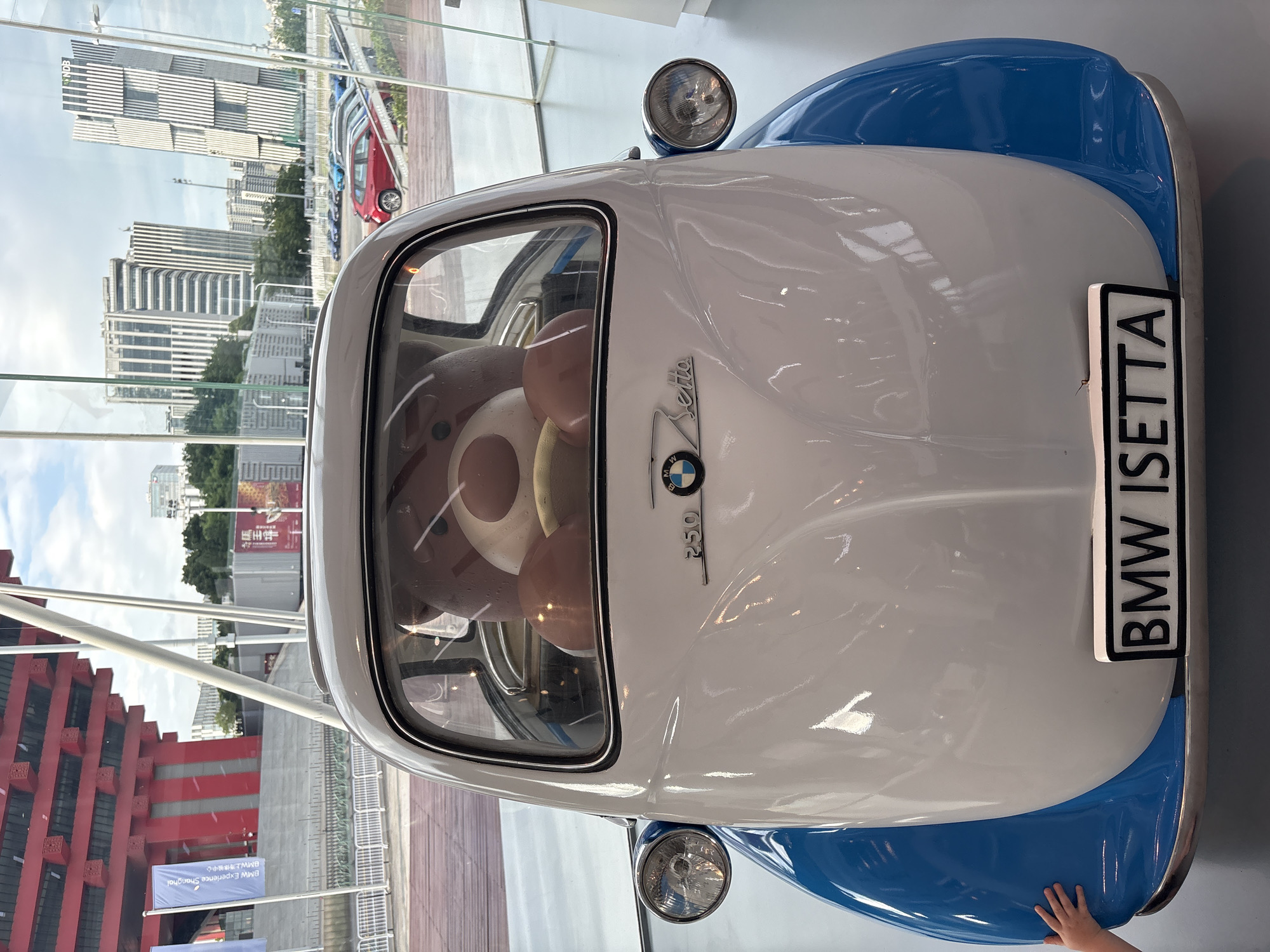 In Pudong, China, a vintage white and blue BMW Isetta car is on display. A large brown teddy bear is seated in the driver's position of the car. The car is parked indoors, with a modern building and skyline visible through a large glass window in the background. A person's hand is visible touching the front fender of the car.