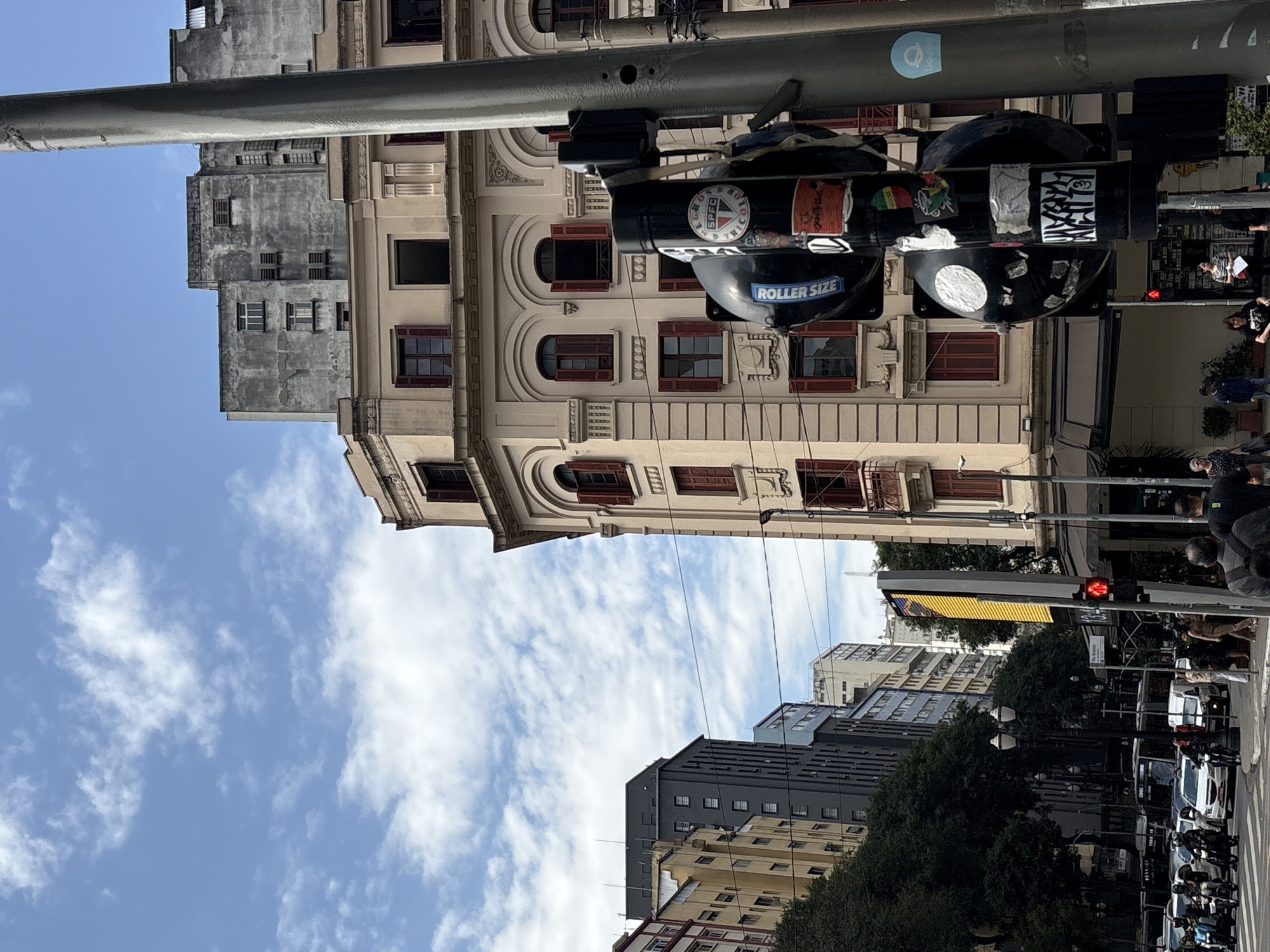The image captures a street scene in São Paulo, Brazil, likely in the downtown area, characterized by its dense urban architecture. Dominating the foreground is a traffic light pole adorned with various stickers. Behind it, an ornate, light-colored building with red shutters on its windows stands prominently. To the left, a cluster of taller, modern buildings, one dark gray and another yellow, create a stark contrast with the older architecture. The sky above is a bright blue with scattered white clouds. The street level shows a crosswalk with pedestrians and a variety of vehicles, including motorcycles, cars, and buses, indicating a busy urban environment. The presence of traffic lights and street signage confirms this is an active intersection within the city.
