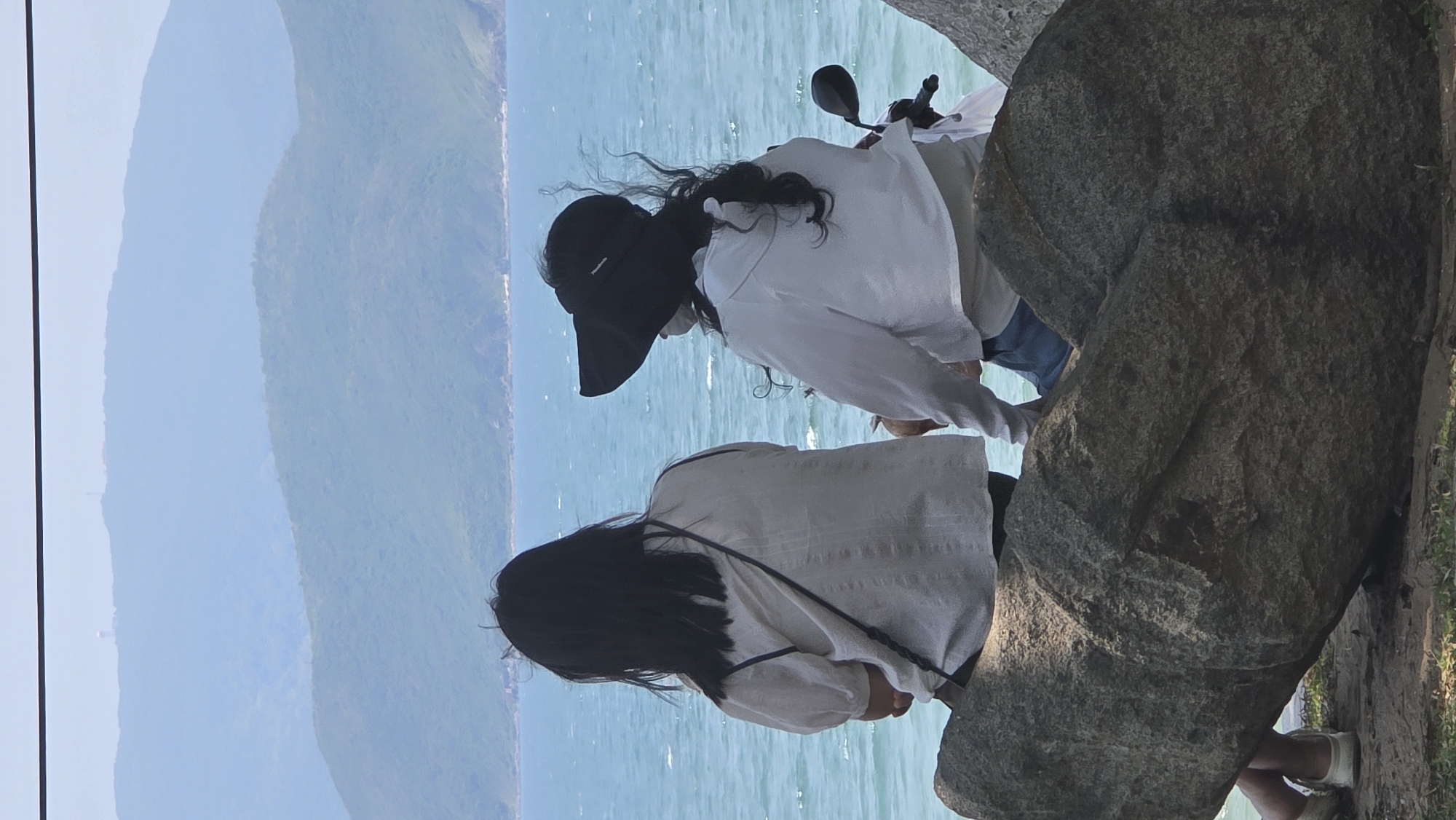Two women stand near a large rock formation overlooking a body of water, likely the sea, with a mountainous backdrop. The woman closest to the viewer has long black hair, wearing a white shirt with a strap of a bag visible across her back, and white shoes. The other woman, partly obscured, wears a black visor and a white shirt and is sitting on a motorcycle. They are located in Nha Trang, Vietnam.