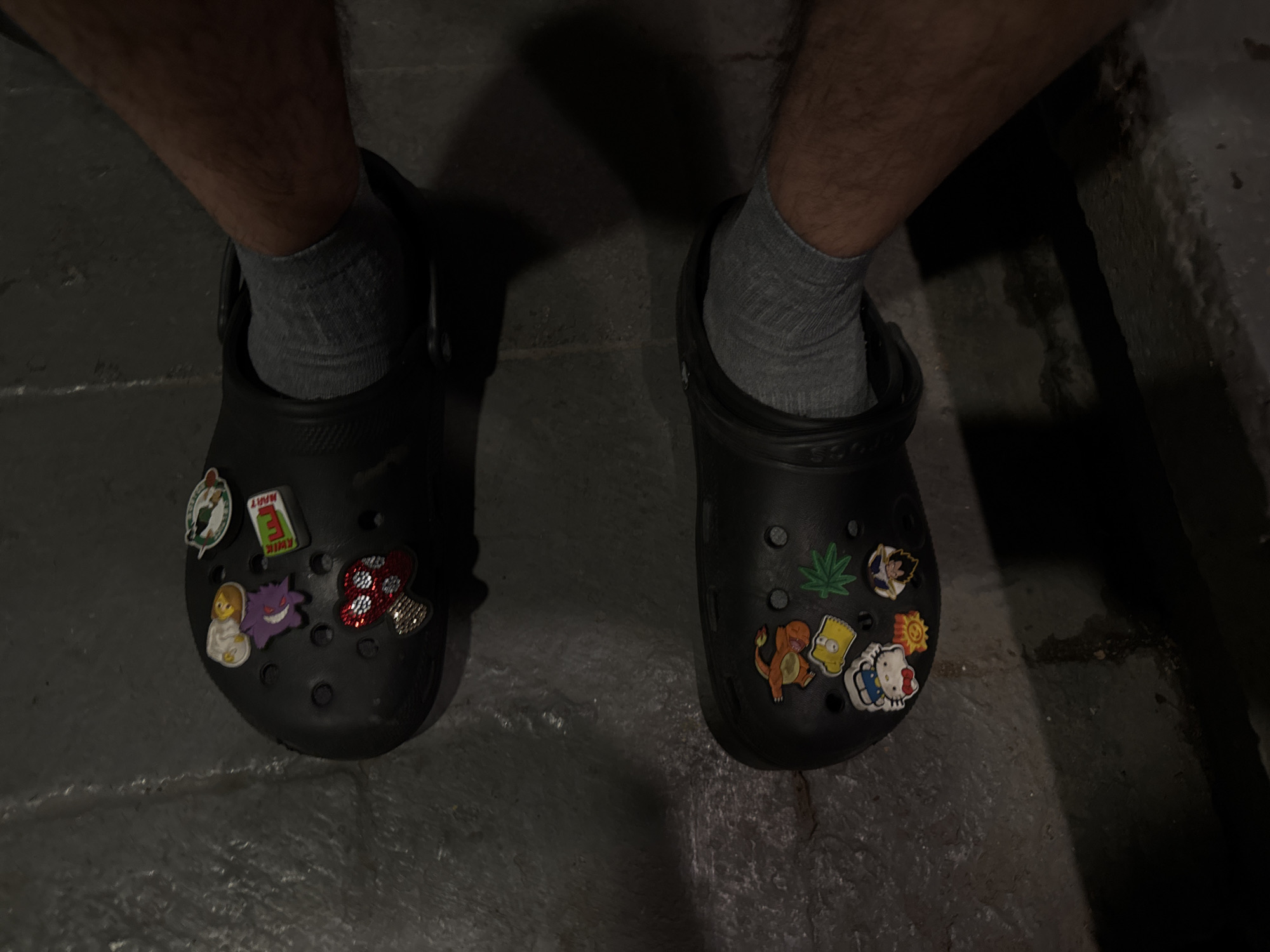 This media file presents a close-up view of a person's legs and feet, clad in black Crocs and grey ribbed ankle socks. The setting appears to be dimly lit, suggesting either an indoor environment or an outdoor area at night. The ground is dark and textured, possibly concrete or tiled.
The Crocs are customized with numerous Jibbitz (charms). On the left Croc, visible charms include a Boston Celtics logo, a "KWIK E MART" sign, a small figure in light clothing (possibly a baby or child character), a purple Gengar Pokémon, and a red mushroom embellished with glitter or rhinestones.
The right Croc displays a green cannabis leaf, a Super Saiyan Goku character from Dragon Ball, an orange Charmander Pokémon, Bart Simpson, a yellow sun icon, and a Hello Kitty charm. The brand name "CROCS" is discernible on the side strap of the footwear. No specific activity beyond wearing the shoes is depicted, nor are any faces or discernible emotions visible. The image is provided in the context of Mexico City, Mexico.