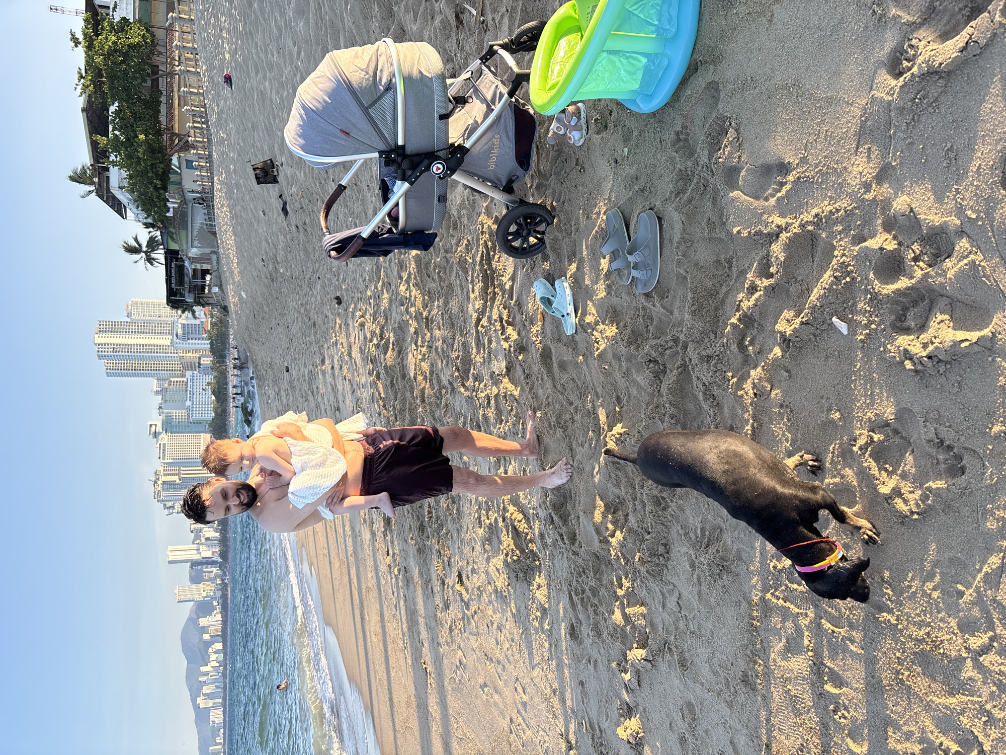 A man is holding a baby wrapped in a white towel on a sandy beach in Nha Trang Ward, Vietnam. The man is shirtless and wearing dark shorts. The baby is facing towards the man's chest. In the background, there is the ocean, a line of buildings, and a mountain. A stroller is parked on the sand near the man and baby. A black dog with a colorful collar is sniffing the sand in the foreground. Several pairs of sandals and a child's inflatable pool are also on the sand. The sun is low in the sky, casting long shadows.