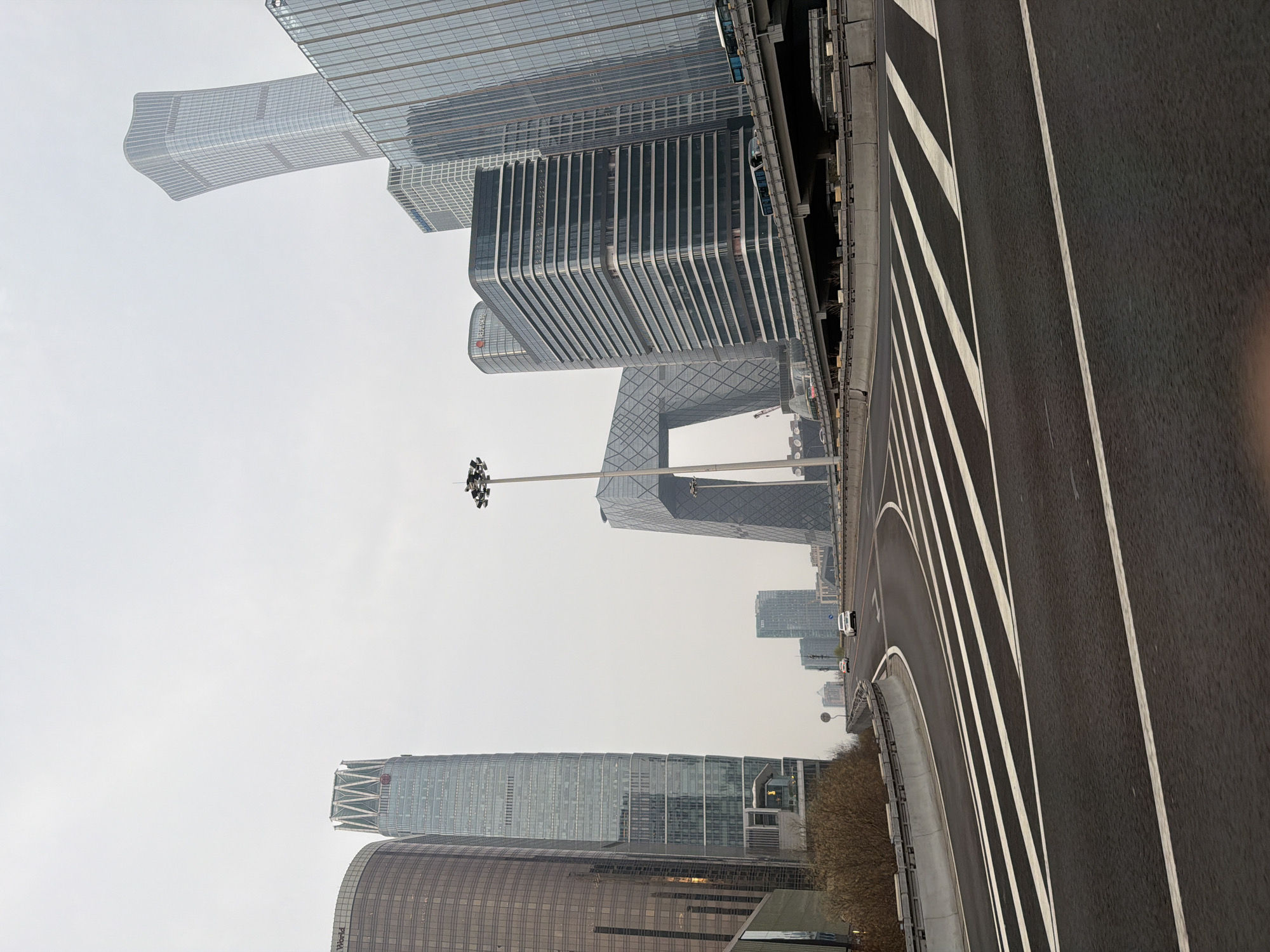 The image shows a street scene in Chaoyang District, Beijing, China, with multiple modern buildings visible against a hazy sky. The CCTV Headquarters building, notable for its unique looped architectural design, is visible. Adjacent to the road, there is a light pole. A roadway is in the foreground, marked with lane lines. A row of trees lines the road.