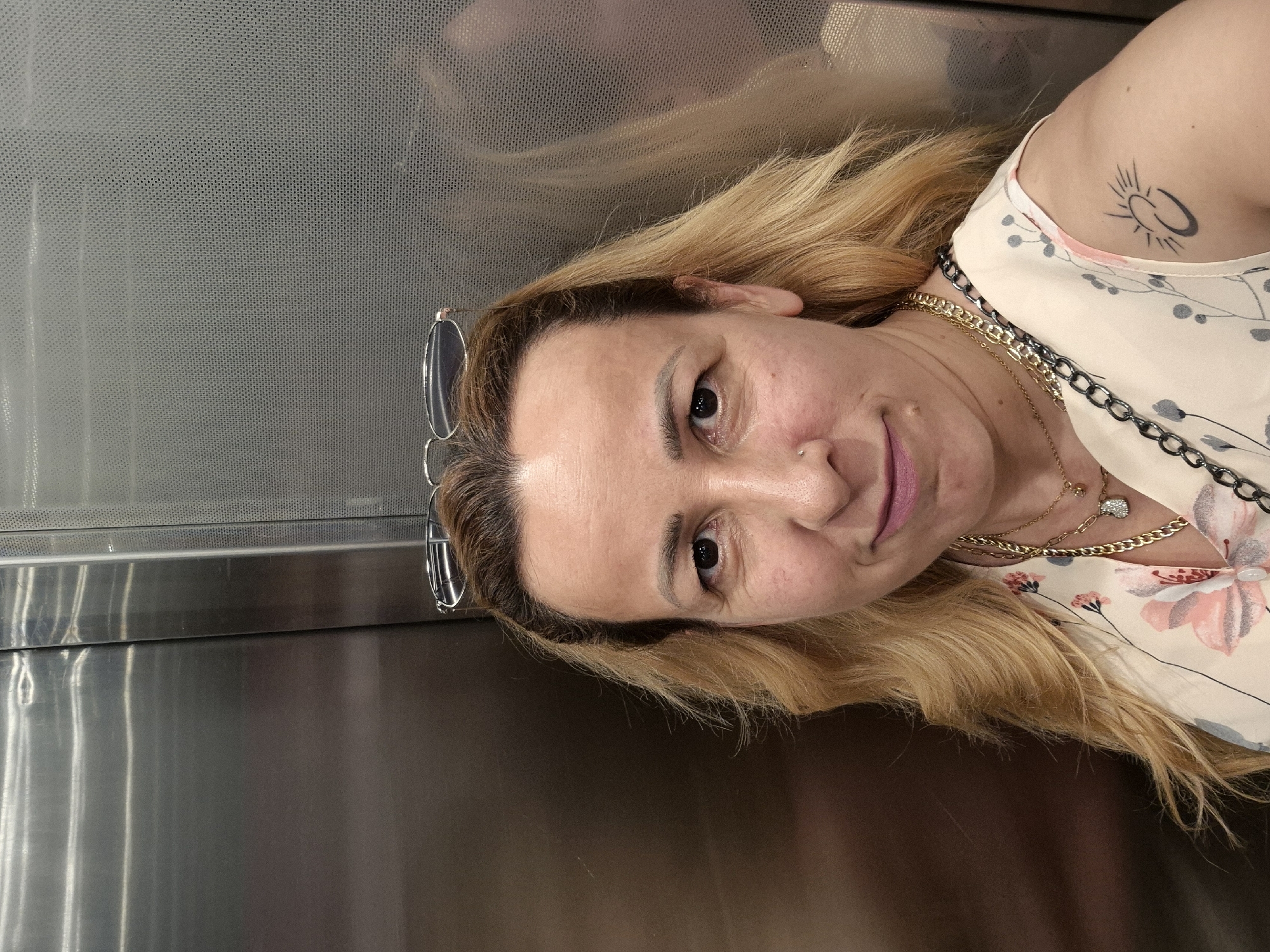 A woman with blonde hair, wearing glasses on top of her head, a floral-patterned shirt, and two necklaces, poses for a selfie in an elevator. The elevator walls are metallic and textured. She has a tattoo of a sun and crescent moon on her left arm.