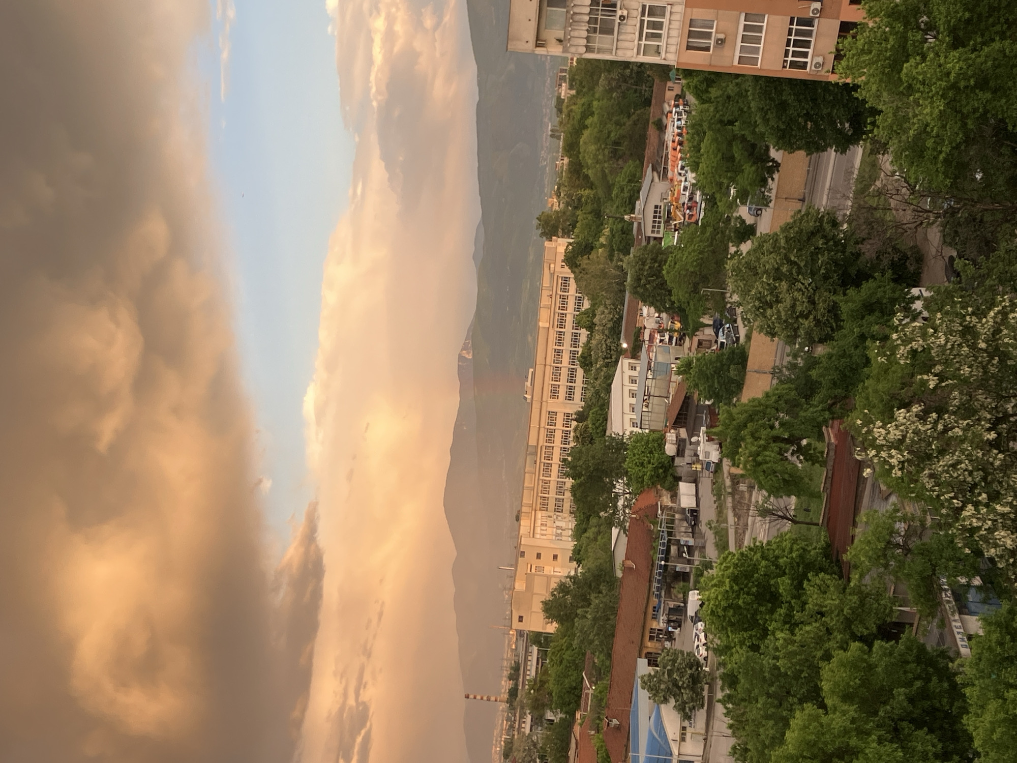 The image shows a cityscape in Plovdiv, Bulgaria, likely taken from an elevated position. In the foreground, there are residential and commercial buildings mixed with lush green trees. A street runs through the middle of the view, with cars parked along the sides. Beyond the cityscape, a mountain range is visible in the background, partially obscured by dramatic storm clouds, colored with orange and yellow hues, suggesting either sunrise or sunset. A tall industrial chimney is visible in the midground. The image is oriented vertically.