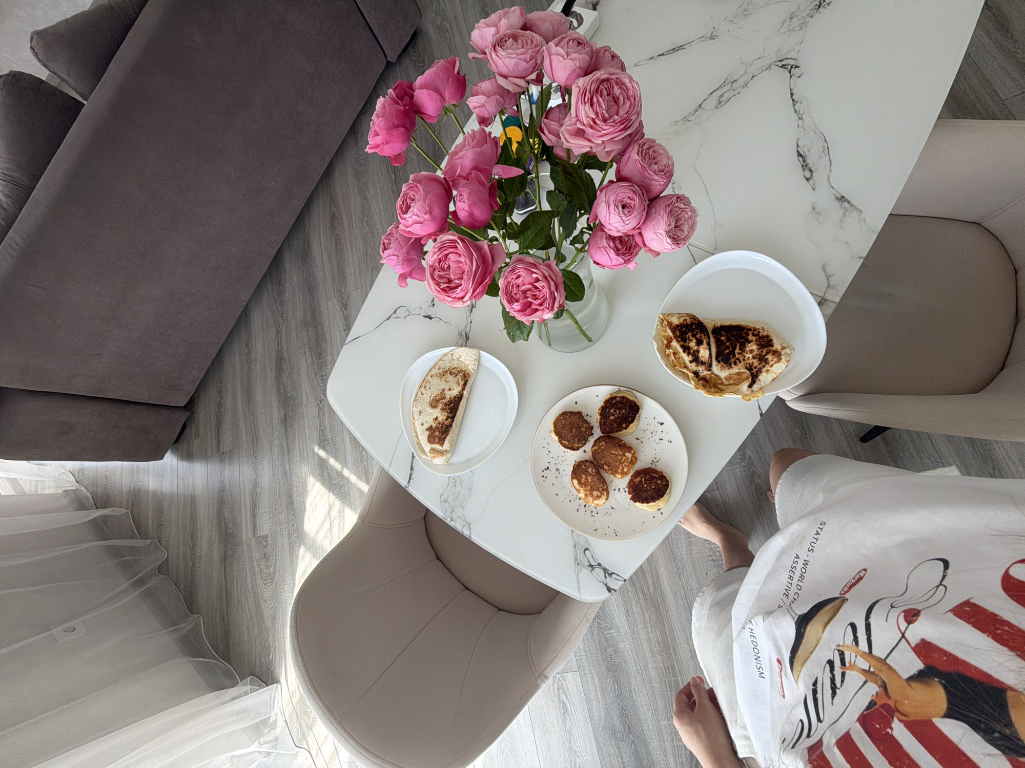 In Malinovka, Russia, a top-down view captures a domestic scene on a marble-patterned table. A bouquet of vibrant pink roses in a clear glass vase dominates the center, its blooms filling the frame with color. To the left of the flowers, a white plate holds a folded, browned flatbread. Below that, another white plate displays five small, round pancakes sprinkled with dark specks. To the right of the roses, a third white plate contains a single folded piece of browned flatbread, appearing to be the same as the one on the left. The table is set on a grey wooden floor, with the legs of a beige upholstered chair visible on the left. In the lower left corner, a person's legs are visible, clad in white shorts with a graphic print featuring a figure and stripes, partially obscuring the floor. The background reveals a section of a grey sofa and a sheer white curtain hanging near a wall-mounted outlet.