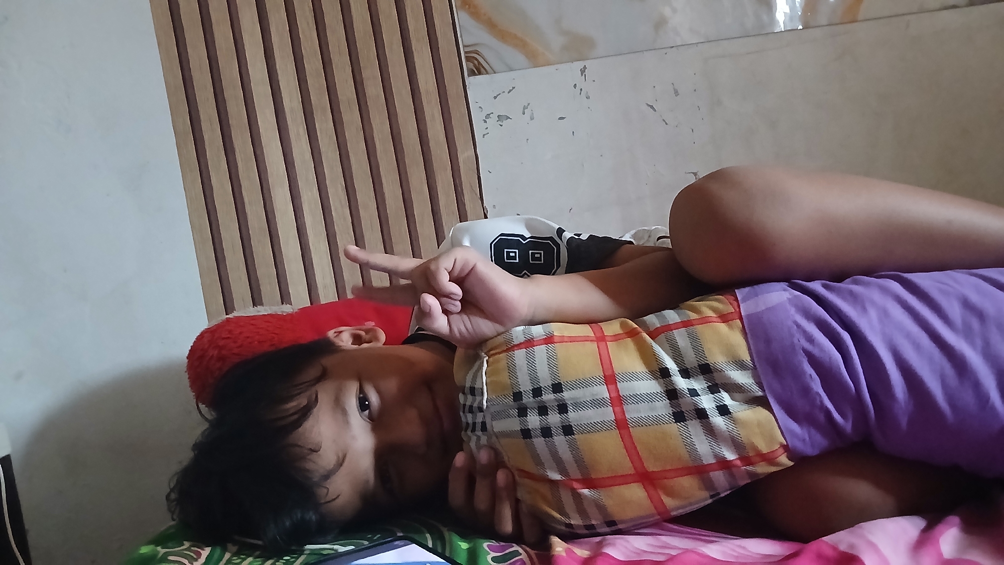 A young Indonesian child is lying on a bed, facing the camera. The child is wearing a yellow and black plaid shirt and a purple blanket is covering their lower body. The child is holding up two fingers in a peace sign. The background consists of a wall with vertical wooden slats and a textured wall with some discoloration. The overall environment appears to be a bedroom in Serah, Indonesia.