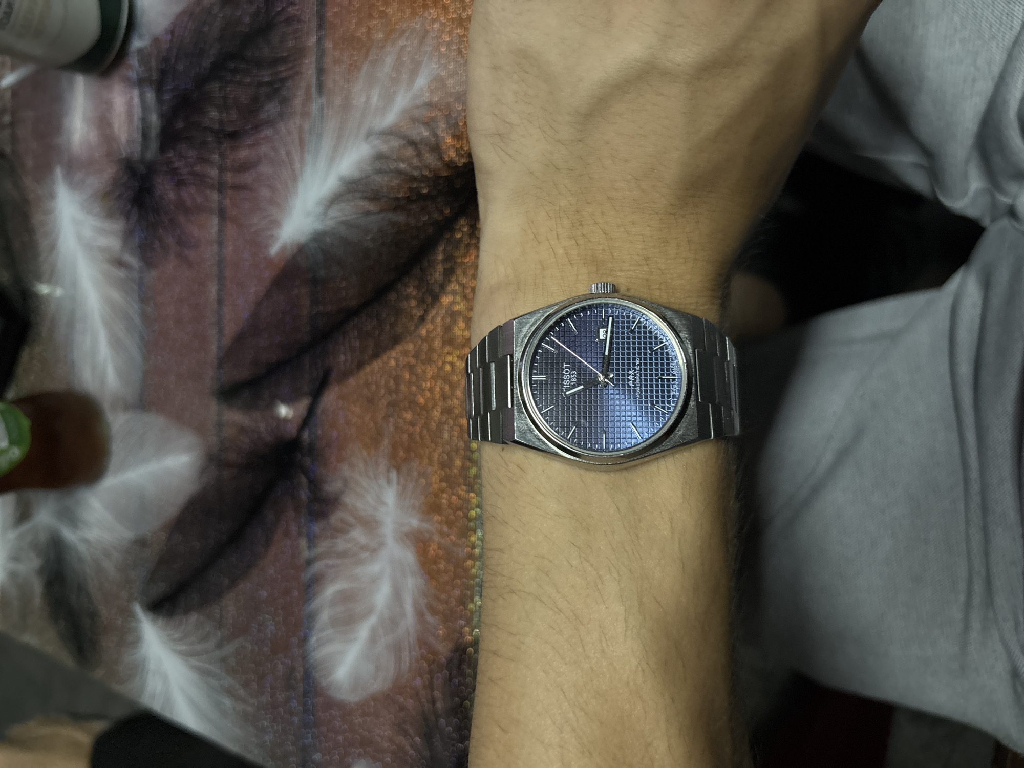 A close-up shot shows a person's wrist wearing a Tissot PRX Powermatic 80 watch. The watch has a silver metal band and a blue dial with a grid pattern. The background is a table with a pattern of white feathers on a brown and purple surface. The overall scene suggests a casual indoor setting.