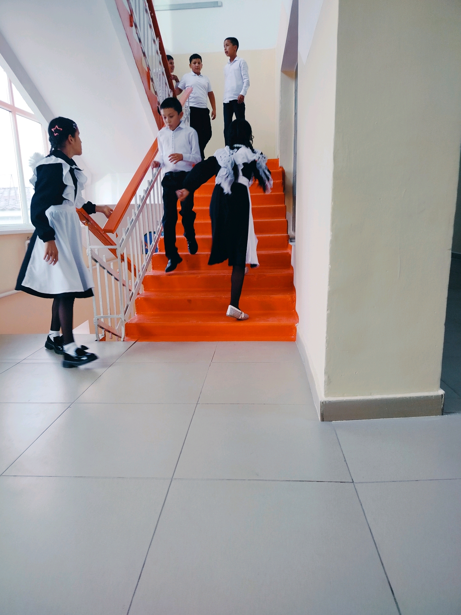 In an indoor school hallway in Kölkent, Kazakhstan, several children in school uniforms are on a bright orange staircase. On the left, a girl with dark hair tied in a braid wears a black and white uniform with a ruffled collar and apron, looking towards the staircase. Two boys in white shirts and black pants are descending the stairs. One boy, further down, is captured mid-stride, while another boy above him is also moving down. Further up the stairs, two more boys in similar uniforms are visible. On the right side of the stairs, a girl in a black and white uniform with a ruffled collar and ribbon is descending, her back to the camera. The hallway has pale yellow walls and a tiled floor. A window is visible on the far left, letting in natural light.