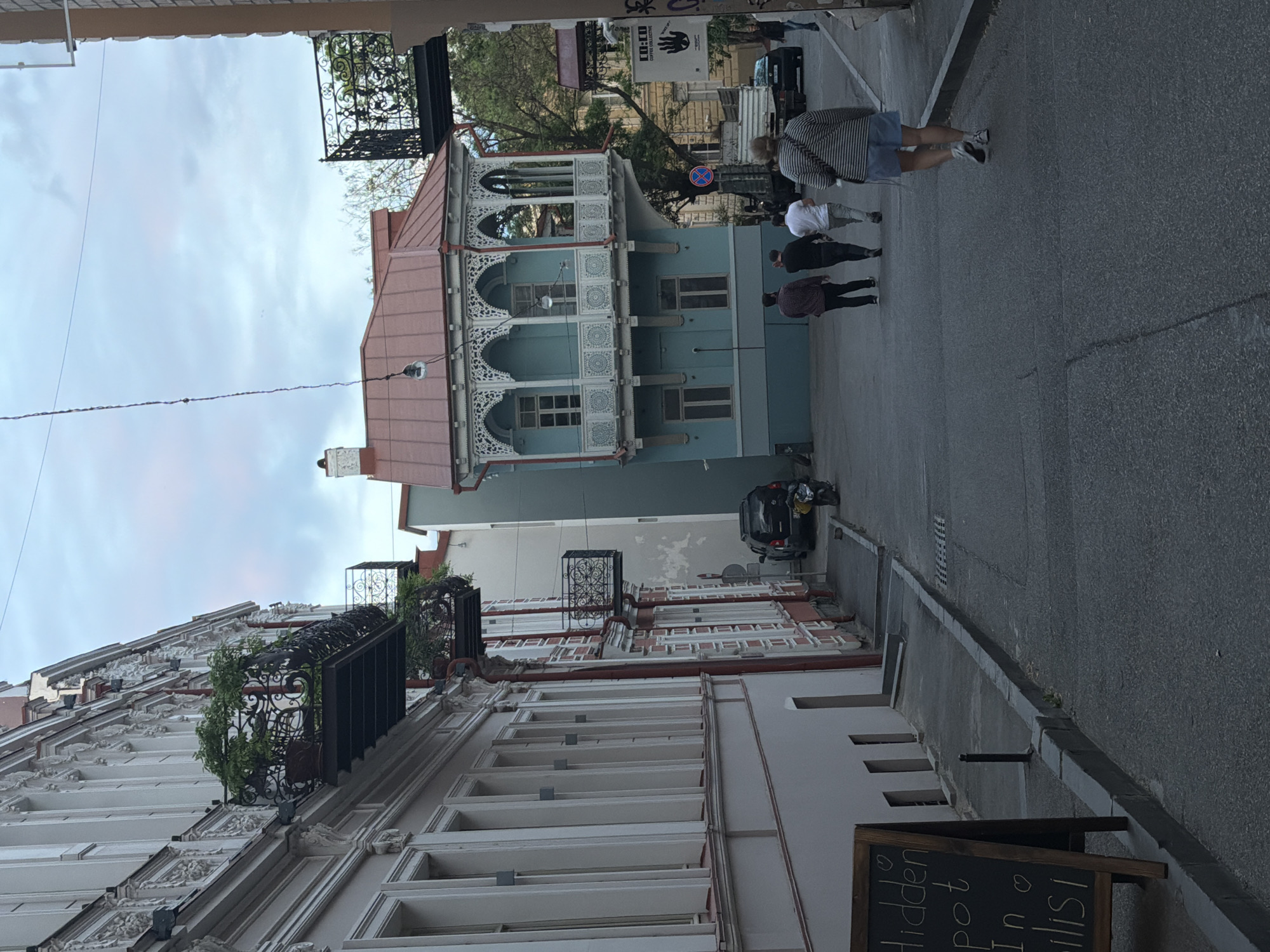 This is a street scene in Tbilisi, Georgia. A narrow street with buildings on either side has pedestrians walking along the right side. The building on the right is white with ornate architectural details, including balconies and ledges. The building across the street is a light blue color with a red roof and decorative white arched balconies. A black car is parked in the street. A chalkboard sign stands on the sidewalk to the right. The sky is overcast.