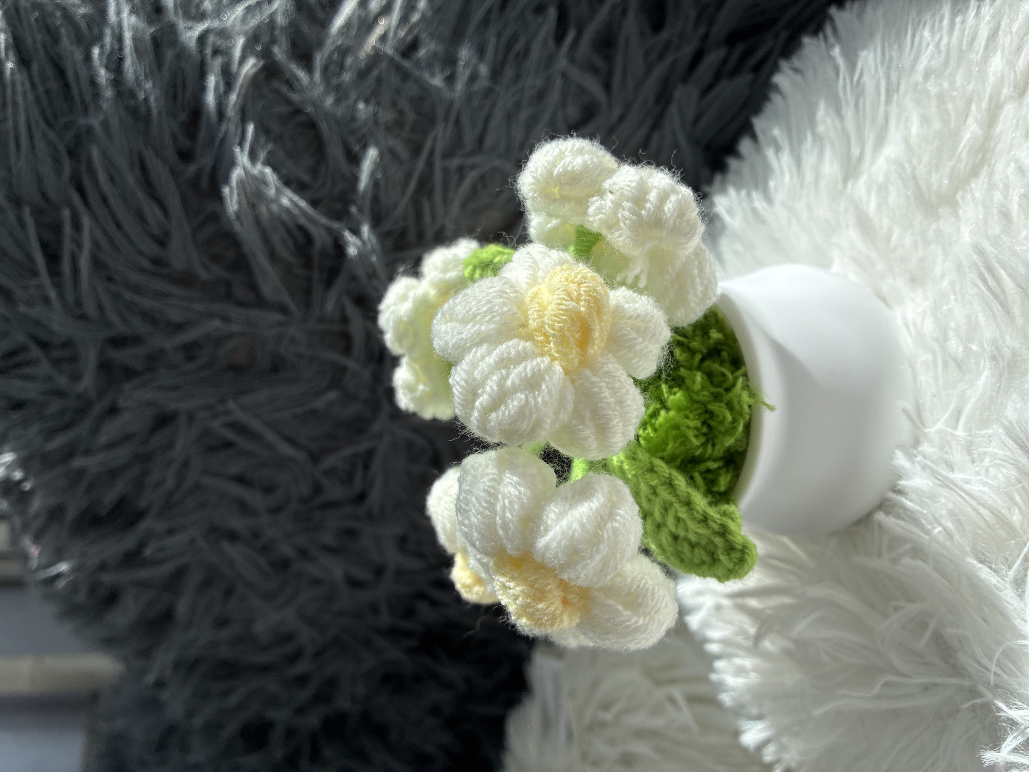 This image features a close-up of crocheted white flowers with yellow centers, nestled in a small white pot. Green crocheted leaves are visible, adding to the floral arrangement. The flowers and pot are positioned on a fluffy white surface, which contrasts with a dark, fuzzy background. The lighting suggests a bright, possibly sunny day, casting shadows and highlighting the textures of the materials. The overall impression is one of delicate craftsmanship and a charming decorative piece. No people, text, or specific location cues are visible beyond the general setting of a still life. The context of Burgas, Bulgaria, is provided externally and is not discernible from the image itself.