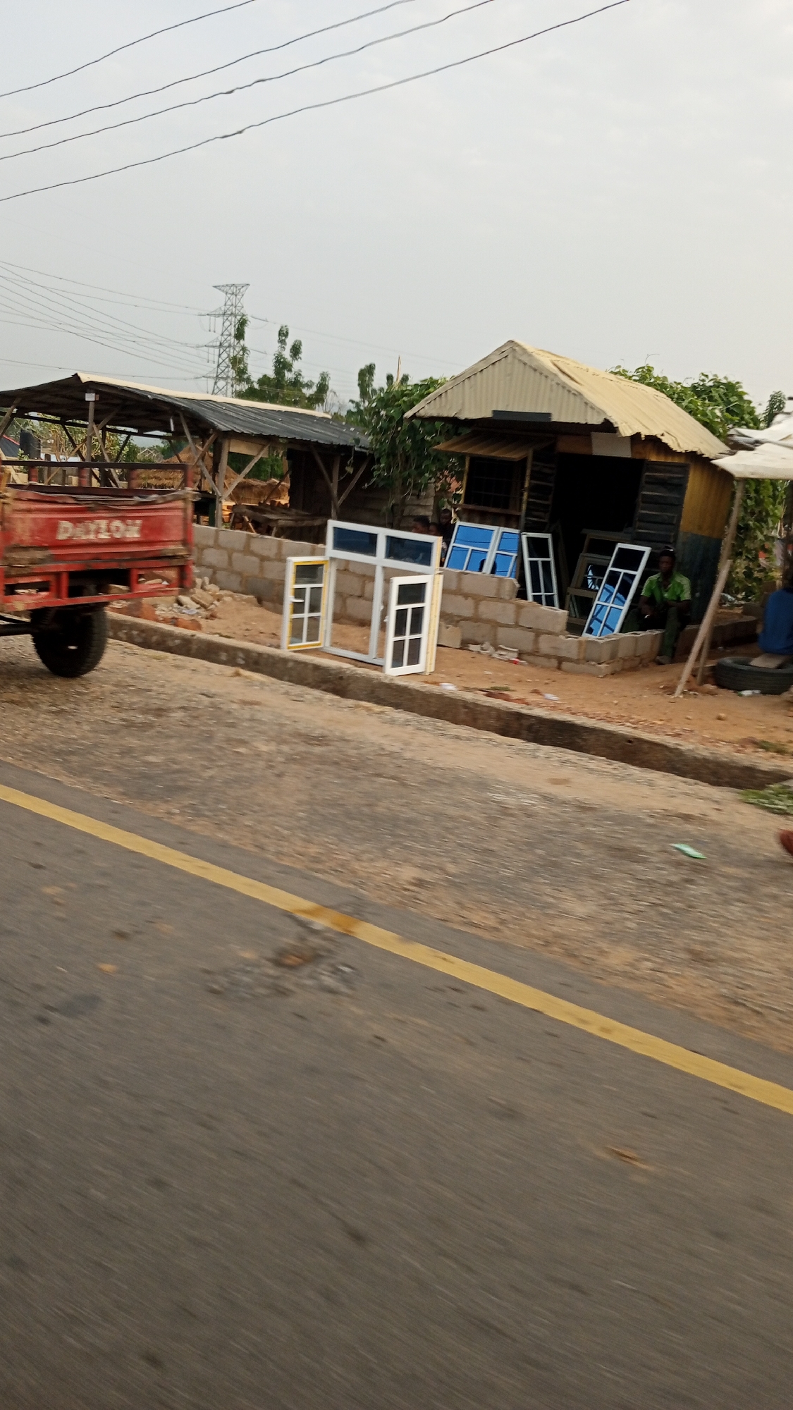 The image shows a roadside scene in Zango-ndaji, Nigeria. A small red trailer is parked on the side of the road, partially obscuring a storefront built from cinder blocks and scrap materials. Several window frames are displayed in front of the storefront, some with blue glass, others with clear or tinted glass. Two people are visible near the storefront; one is sitting, and one is partially obscured. A yellow line is painted on the asphalt road. Power lines and a transmission tower are visible in the background.