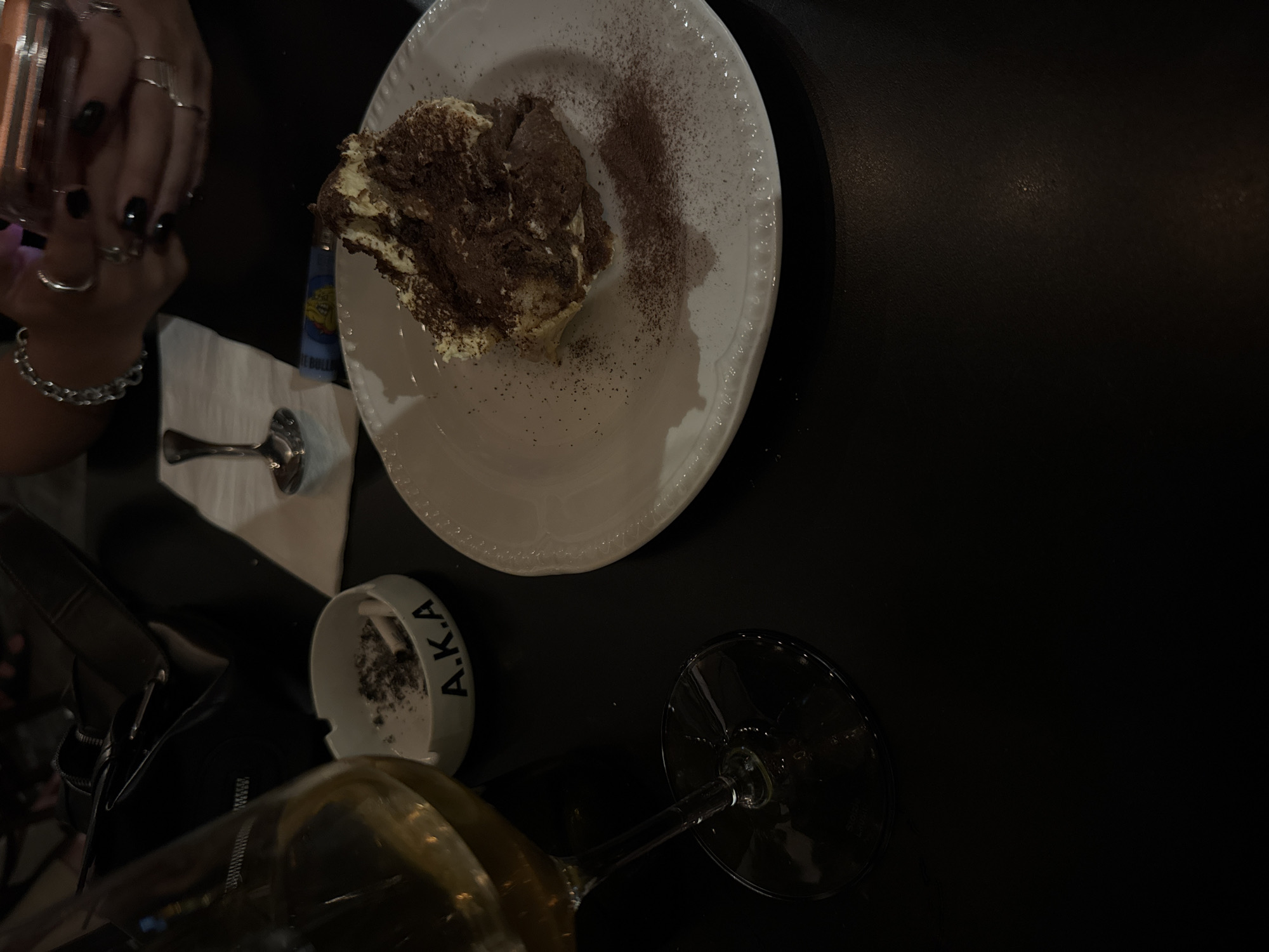 The image depicts a table setting, likely indoors. A portion of a person's hands is visible in the upper left, adorned with multiple rings and a bracelet. A white plate holds a slice of tiramisu with cocoa powder dusting. A spoon and what appears to be a lighter are next to the plate. An ashtray with the inscription "A.K.A" containing cigarette ashes sits near the bottom. The base of a wine glass is also visible at the bottom.
Given the "A.K.A" inscription on the ashtray, it can be inferred that the image was taken at the establishment known as A.K.A in Tel Aviv, Israel.