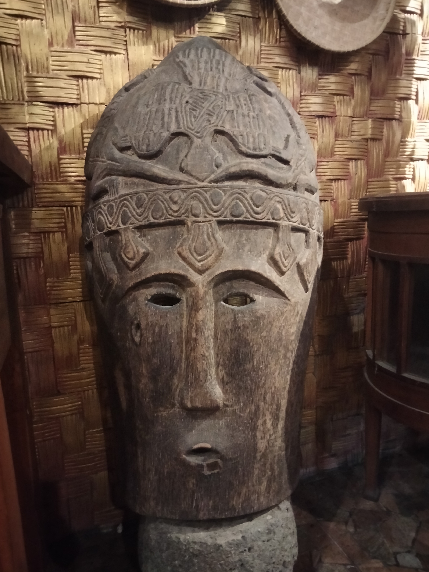 A close-up, eye-level shot shows a weathered, wooden, carved mask displayed in what appears to be an indoor setting with woven bamboo walls. The mask, which is the central subject, is dark brown with intricate carvings around the forehead and above the eyes. The facial features, including the nose, eyes, and mouth, are rendered in a stylized manner. The mask rests on a rough, grey, circular base. To the right of the mask, a portion of a wooden display cabinet with glass panels is visible. The background consists of the textured woven bamboo wall. The lighting is dim, casting shadows on the mask and the surrounding environment. The location is identified as Sukodono, Indonesia.