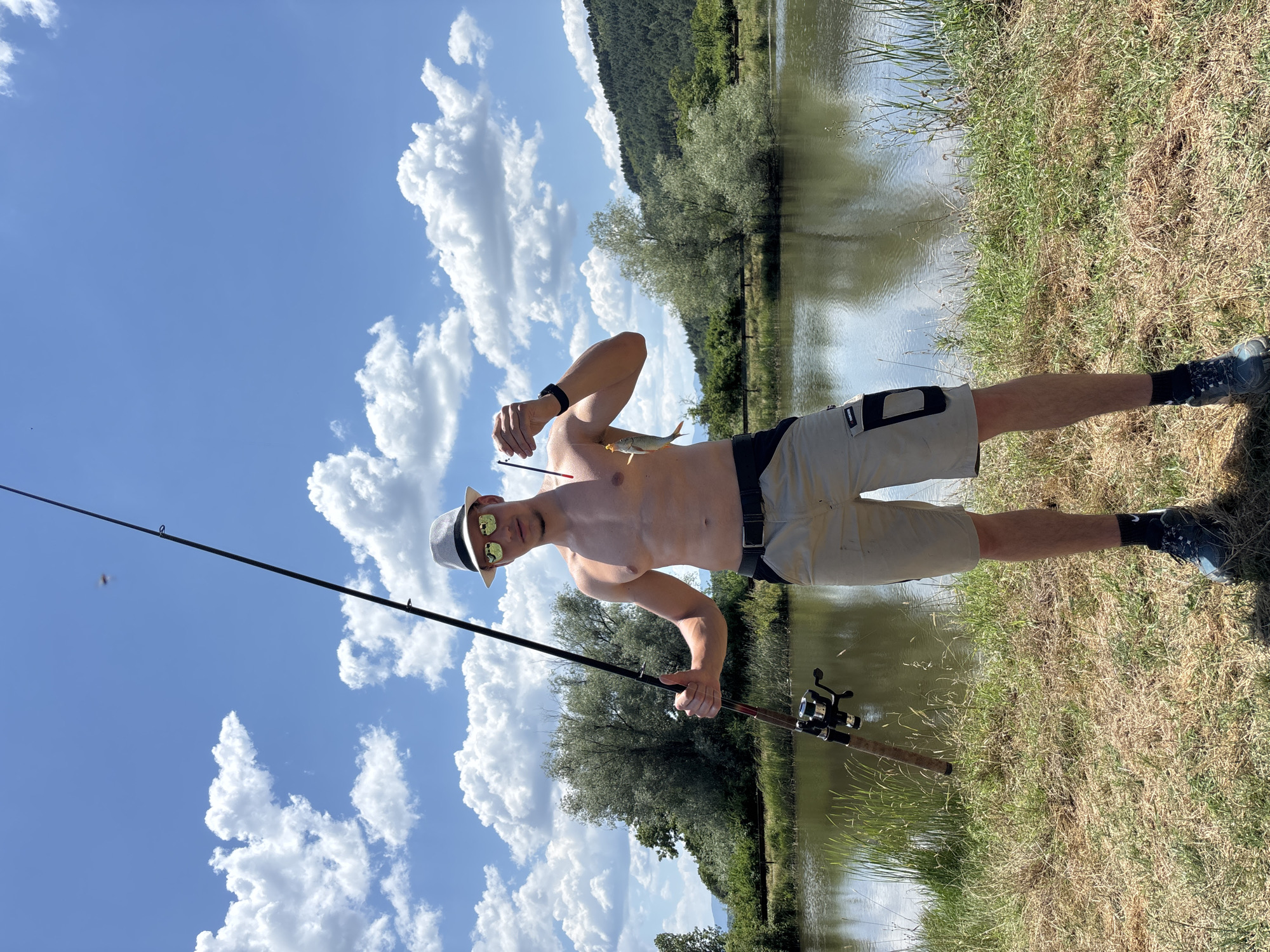 A shirtless man with sunglasses and a white hat stands outdoors by a body of water, holding a fishing rod and a small fish. He is wearing khaki shorts and dark hiking boots. The man is looking at the camera, with his left arm extended and his right hand holding up the fish. The background features a calm body of water, lush green trees, and a hillside covered in pine trees. The sky is a vibrant blue with scattered white clouds. The scene appears to be a fishing spot in Velikdenche, Bulgaria.