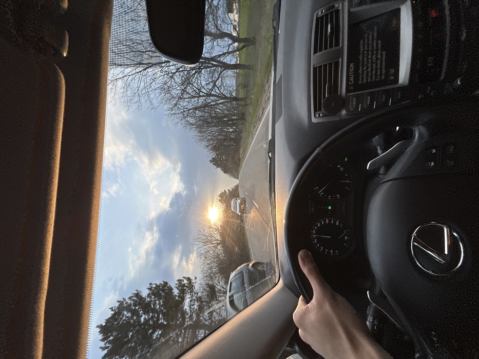 A first-person perspective image captures the interior of a Lexus vehicle and a rural road scene at sunset, ostensibly in Grigorevo, Bulgaria. A driver's left hand is positioned on the black leather steering wheel. The vehicle's instrument cluster indicates operation, and the central infotainment screen displays a "CAUTION" message related to traffic regulations. Directly ahead, the low setting sun causes significant glare and lens flare across the windshield, which exhibits multiple small streaks. The two-lane asphalt road stretches forward, marked with a solid white shoulder line. An oncoming white vehicle is visible in the distant lane. To the immediate left, a dark-colored SUV is partially visible, in the process of passing or having just passed the viewer's vehicle. The roadside environment features rows of bare deciduous trees and scattered evergreens under a partly cloudy sky, characteristic of a late winter or early spring evening.
