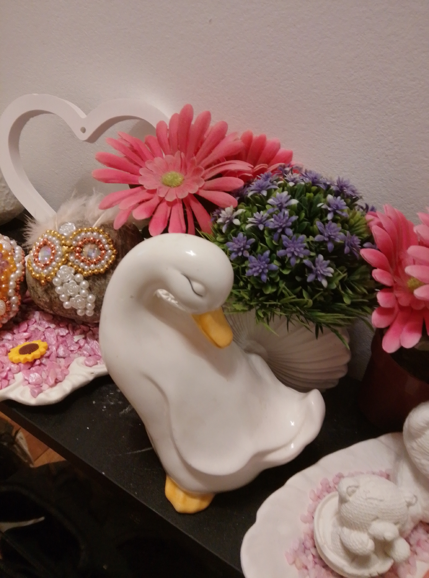 The image displays a collection of decorative items on a dark shelf. In the foreground, a white ceramic duck with a yellow beak is positioned to the right. To the left of the duck is a white ceramic dish with small pink gravel and a yellow sunflower-shaped charm. Behind the duck, a potted plant with small purple flowers sits in a white, shell-shaped planter. Pink and white flowers are arranged behind the plant. To the far left, a white heart-shaped decoration is visible. Further to the left, a decorative sphere adorned with beads and pearls is partially visible, alongside some white feathers. The background is a plain, textured wall. The scene appears to be indoors, possibly a home or shop display. The image was taken in Wedel, Germany.