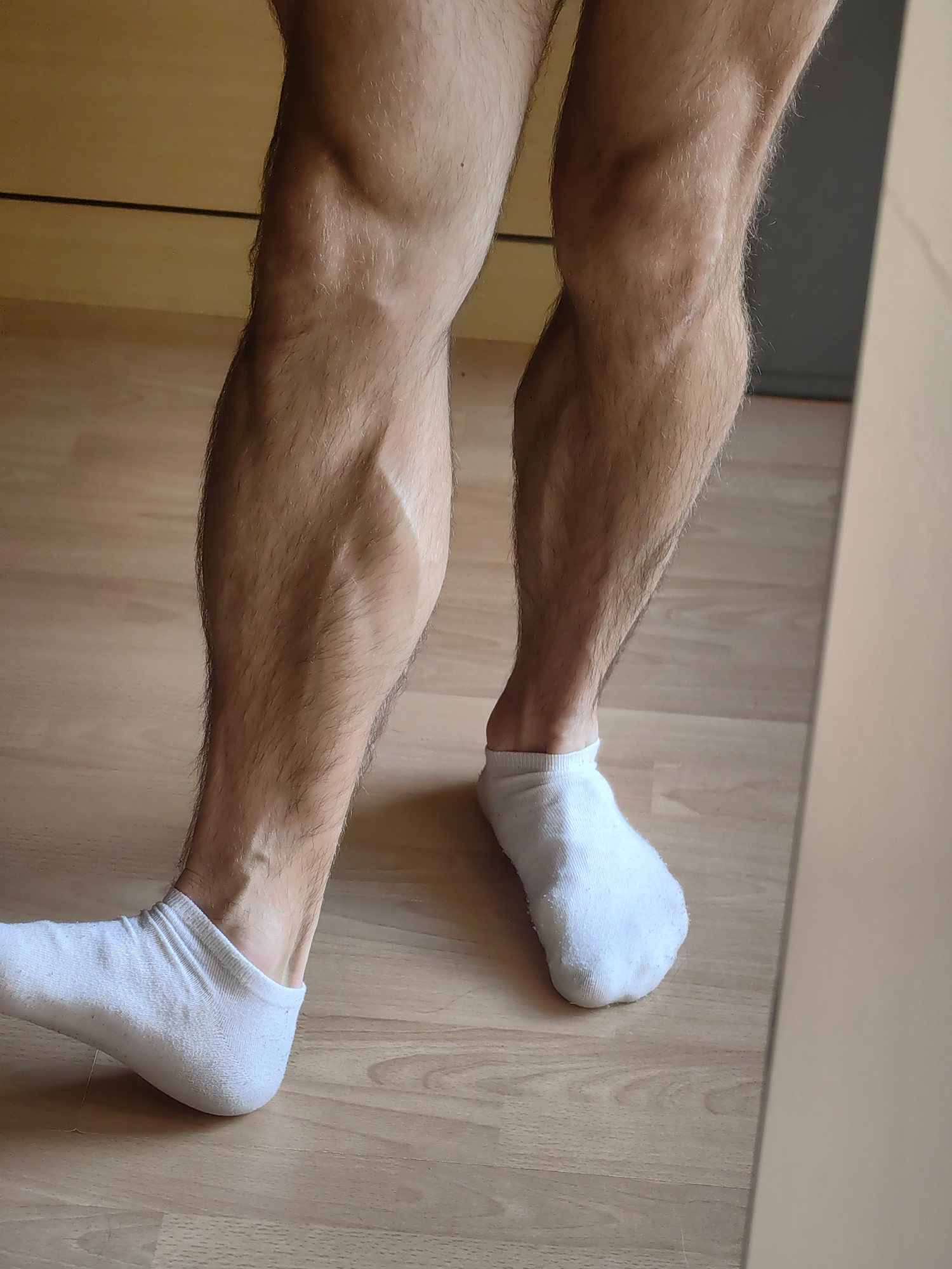 The image shows a close-up view of a person's lower legs and feet, which are clad in white ankle socks. The legs are muscular and hairy, with prominent veins visible on the calves. The person is standing on a light brown wooden floor. In the background, there are hints of wooden cabinetry or furniture. The lighting suggests an indoor environment, possibly a domestic setting. There is no indication of the city of Varna, Bulgaria, or any urban elements within the image.