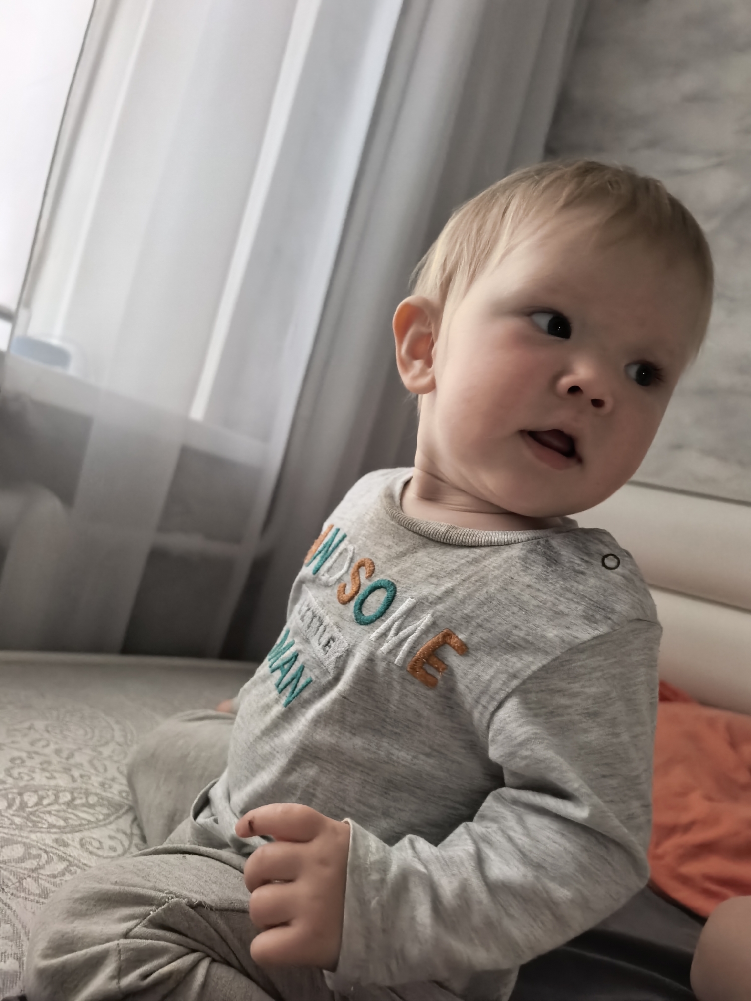 A blonde toddler with fair skin and light brown eyes is seated on a bed. The toddler is wearing a grey long-sleeved shirt with embroidered text that reads "HANDSOME LITTLE MAN" and grey sweatpants. The toddler is looking to the right of the frame with their mouth slightly open. The background consists of a sheer curtain on the left, a textured wall or surface behind the toddler, and part of an orange pillow on the right. The setting appears to be a bedroom. The image does not contain any specific information to indicate it is located in Sterlitamak, Russia.
