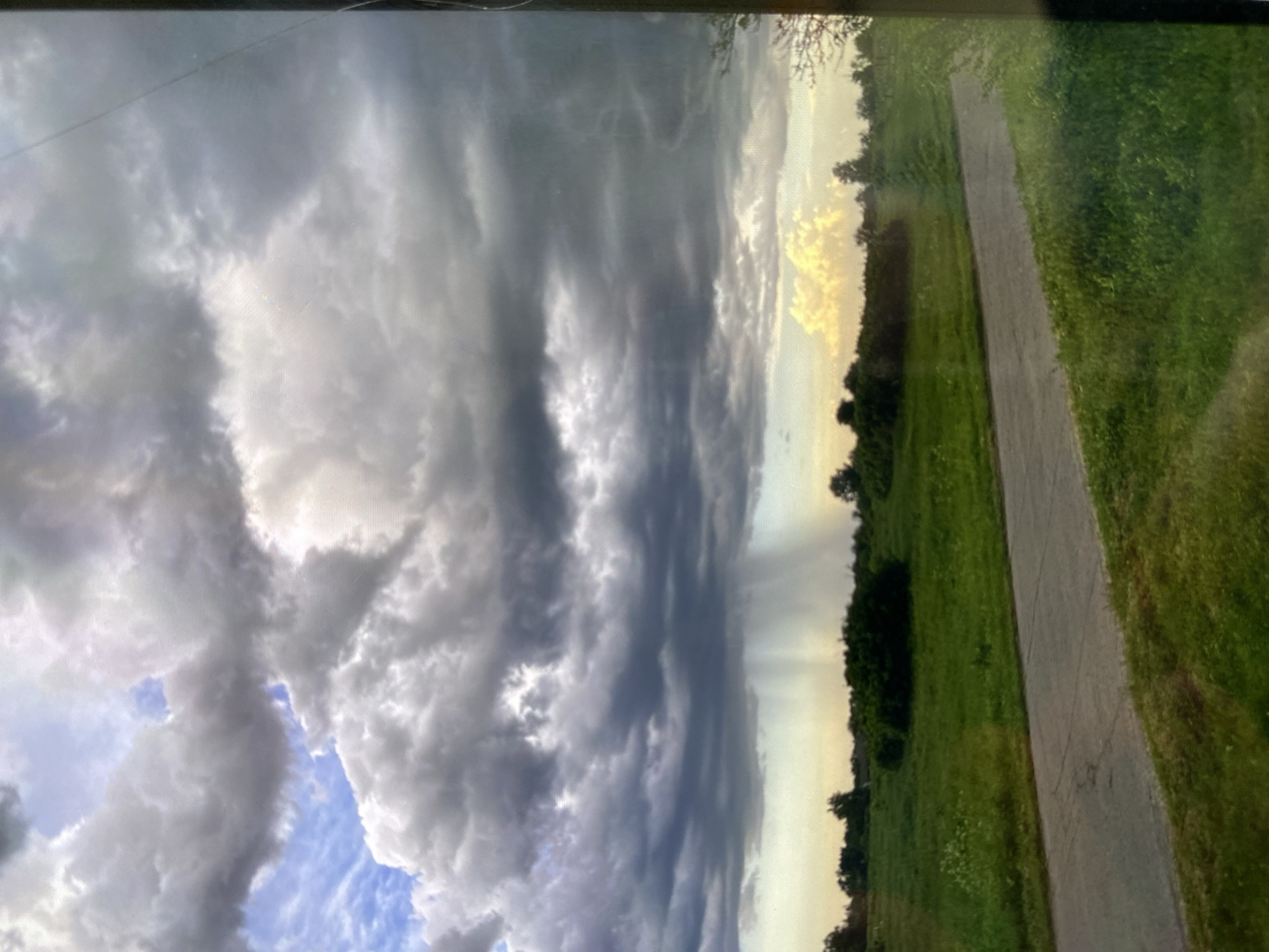 The image depicts a rural landscape under a dramatic sky. Heavy, dark clouds dominate the upper portion of the frame, with a distinct shaft of rain falling in the distance. The ground below is covered in lush green grass, with scattered trees and bushes dotting the field. A paved road or path cuts through the scene from the foreground, leading towards the horizon. The lighting suggests either early morning or late afternoon, with a warm glow on the right side of the sky where a patch of lighter clouds is visible. No people or animals are present in the image. The scene appears calm and natural, despite the impending or ongoing rain.