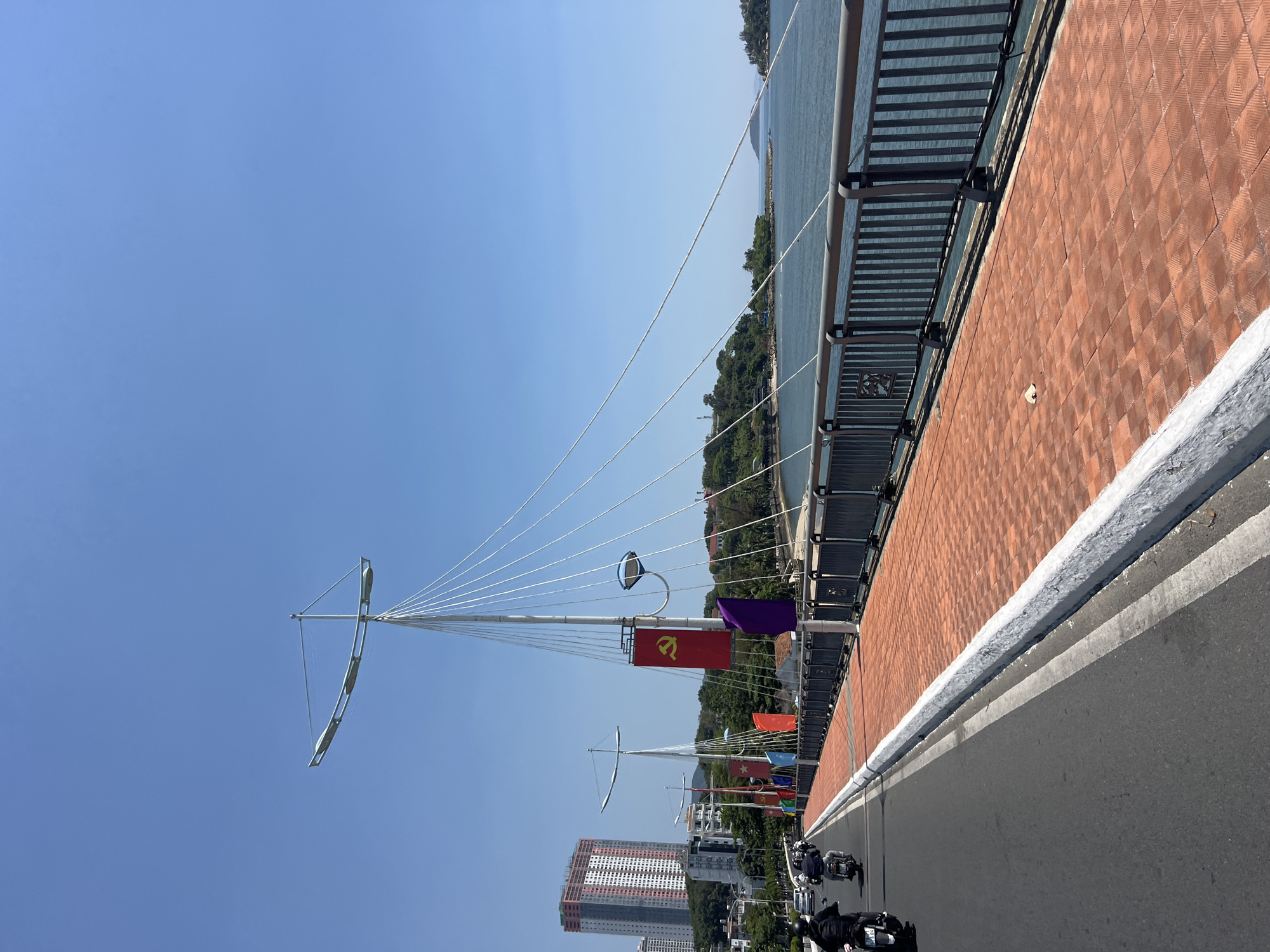 The image depicts a bridge in Nha Trang, Vietnam, under a clear blue sky. The bridge features a brick-patterned pedestrian walkway along one side and a gray asphalt roadway. Metal railings run along the edge overlooking a body of water and a tree-lined area in the distance. Decorative masts with thin cables extend upwards, and several flags, including one with a hammer and sickle symbol, are attached to the masts. A tall building is visible in the background. Several motorcycles are on the roadway.