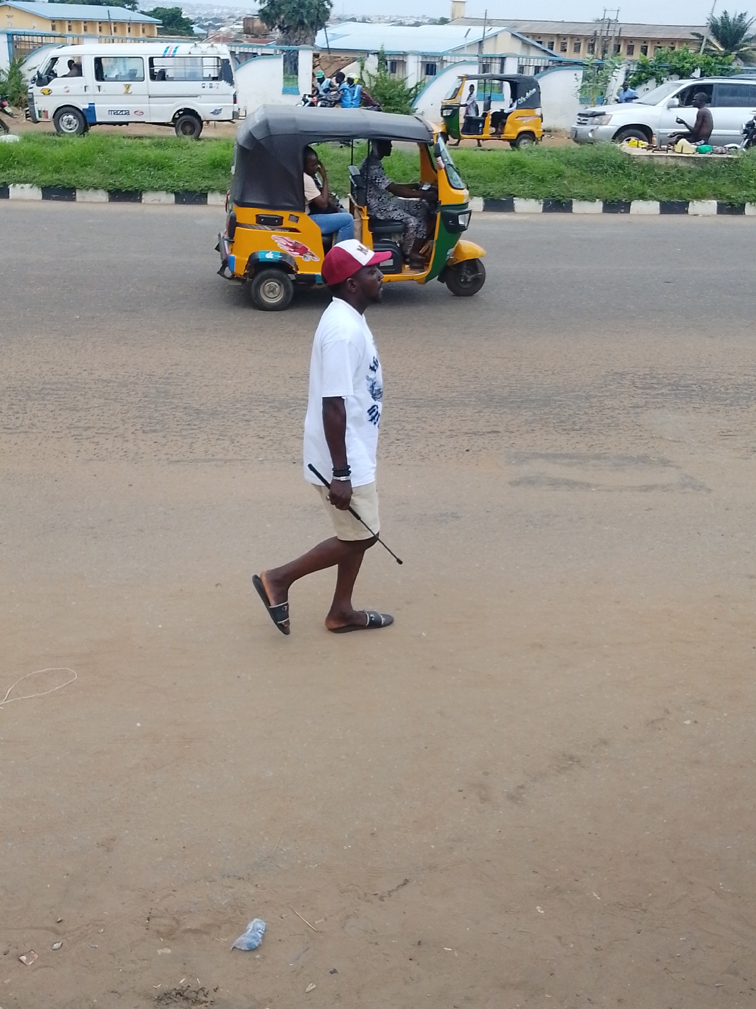 In Oja Oba, Oko Erin, Nigeria, a man walks on a dirt road, wearing a white t-shirt, beige shorts, and black and white sandals. He is holding a black stick in his right hand and has a red and white baseball cap on his head. Behind him, a yellow and black auto-rickshaw is on the road, with two people inside. To the left of the auto-rickshaw, a white van with blue stripes is parked. Further in the background, a silver SUV is parked on the side of the road, and a person is sitting near it. Buildings and trees are visible in the far background. The ground is covered in dirt and small debris.