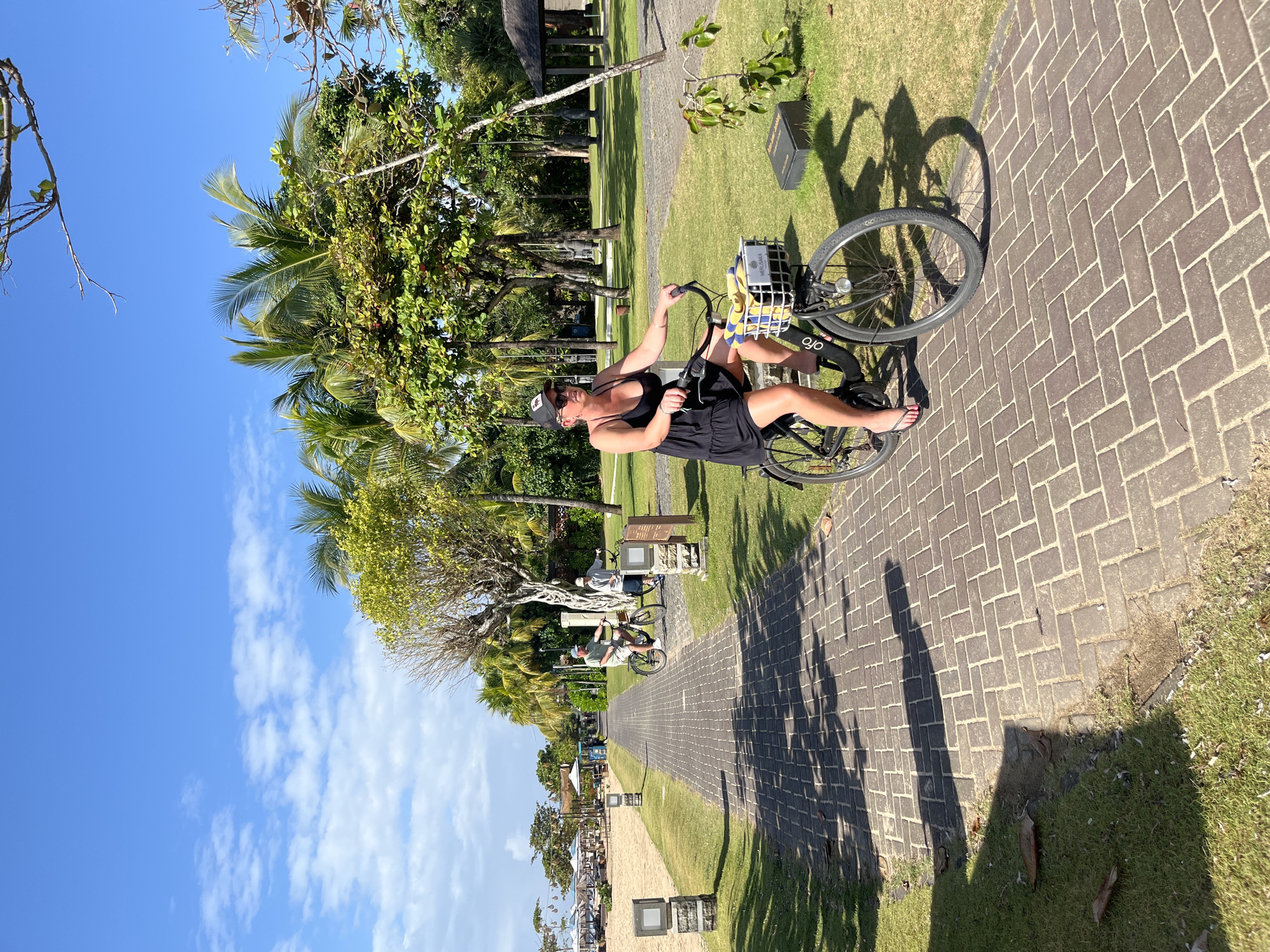 In Kuta Selatan, Indonesia, a woman with blonde hair and sunglasses is riding a bicycle on a paved pathway. She is wearing a black dress and flip-flops. The bicycle has a basket in front containing yellow and blue striped towels. She is on the right side of the pathway, which is made of brick pavers. To her left is a grassy area with some trees, including palm trees, and to the far left is a sandy beach with beach chairs and umbrellas. Further down the pathway, two other individuals are riding bicycles. The sky is blue with scattered white clouds. The scene appears to be a resort or park setting, with lush vegetation and tropical elements.