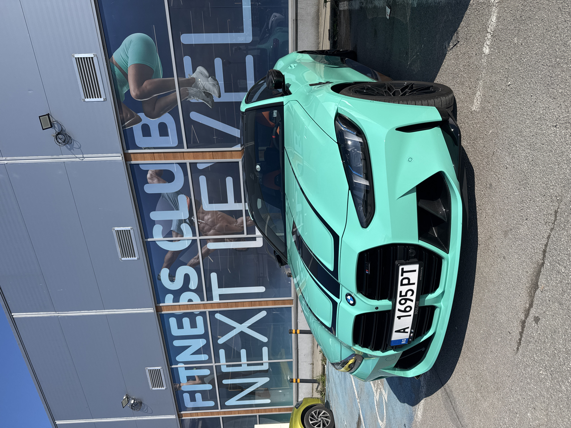 A mint-green BMW car with two black racing stripes is parked on a gray asphalt surface next to a building in Burgas, Bulgaria. The building appears to be a fitness center with large glass panels displaying the words "FITNESS CLUB NEXT LEVEL". The car has a Bulgarian license plate "A 1695 PT". Part of another yellow vehicle is visible in the bottom left corner of the image. The sky is clear and blue.