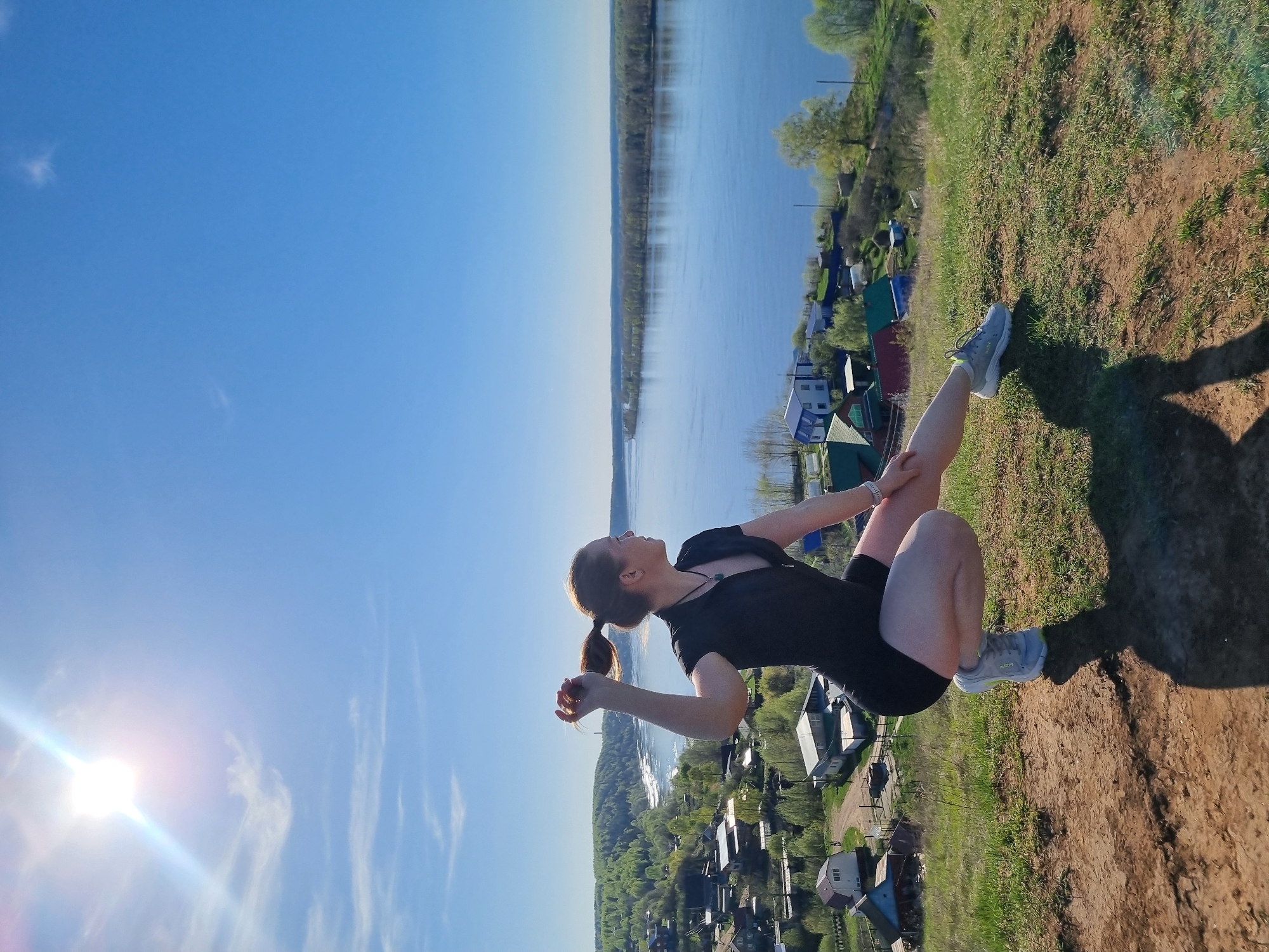A woman in athletic attire is sitting on a grassy hill overlooking a river and houses. The river stretches into the distance with tree-lined banks. The houses, of various colors, are nestled among the trees on the near bank of the river. The sky is a clear blue with the sun shining brightly, casting a glare on the lower left of the image. The woman is wearing a black top and shorts, and gray sneakers. Her hair is pulled back into a ponytail. Her legs are bent and one hand is resting on her knee. The background suggests a natural environment close to a settlement. The location is Ольховка, Russia.