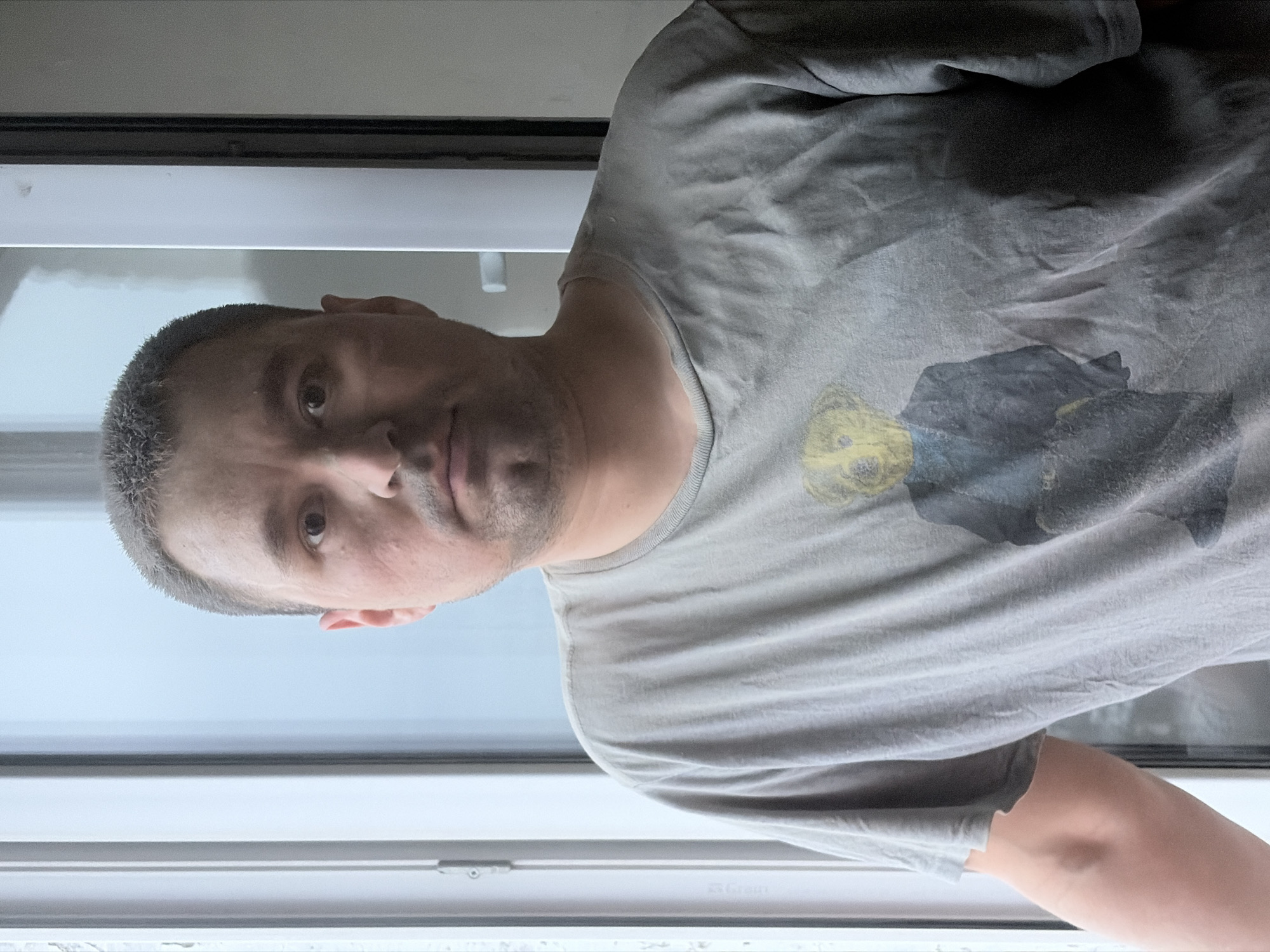 A man is standing in front of a window, looking at the camera. He is wearing a gray t-shirt with a faded graphic of a cartoon bear on it. The man has short, graying hair and stubble. He is indoors, and the background shows a blurred view through the window. The location is potentially Izhevsk, Russia, but this cannot be verified from the image.
