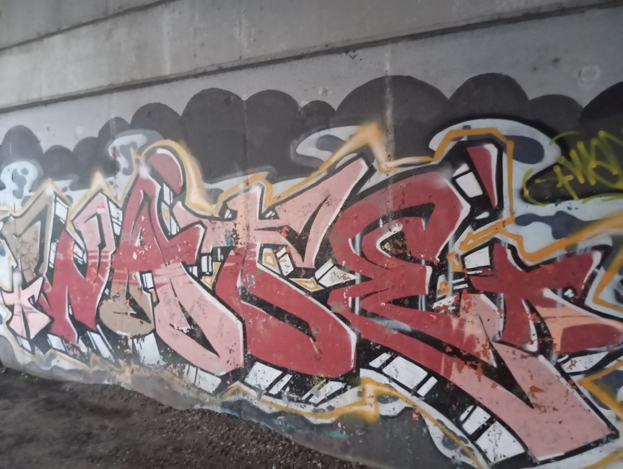 The image shows graffiti on a concrete wall. The graffiti is colorful, with predominantly red, pink, and yellow hues. It features bold, stylized lettering and abstract shapes. The concrete wall appears to be part of an underpass or bridge, with a rough texture. The ground in the foreground is covered with gravel and dirt. The scene is an outdoor urban environment.