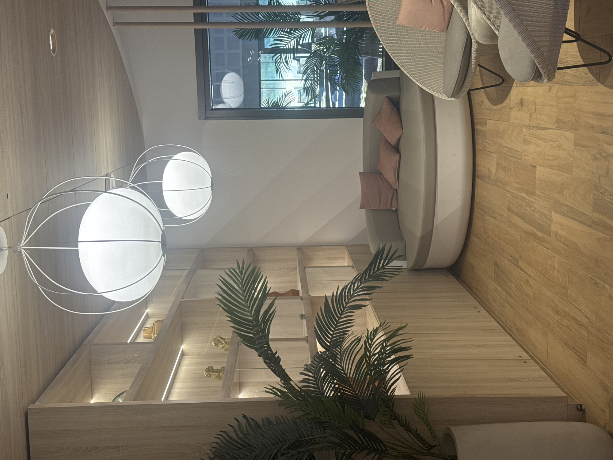 The image presents an interior space, likely a waiting or seating area within a building in Marcq-en-Barœul, France. The perspective is from a lower angle, capturing a partial view of the room's features.
On the left, there is a light-wood paneled wall with built-in shelving, some of which are illuminated. The shelves hold decorative items, including small figurines and what appears to be a perfume bottle.
Above the shelves, a modern, multi-bulb lighting fixture with spherical white shades hangs suspended. This complements the contemporary design of the space.
Moving towards the center, a potted plant with large, green fronds stands, partially obscuring the view of the shelving unit.
The upper right quadrant of the image showcases seating arrangements. A curved sofa with pale upholstery and pink cushions is visible, accompanied by two chairs made of wicker or a similar woven material.
A large window is present at the top, through which one can see exterior elements, including palm trees and architectural structures. The window suggests a tropical or subtropical theme, possibly reflected in the interior decor.
The floor is made of light-colored wood, adding warmth to the overall ambiance.