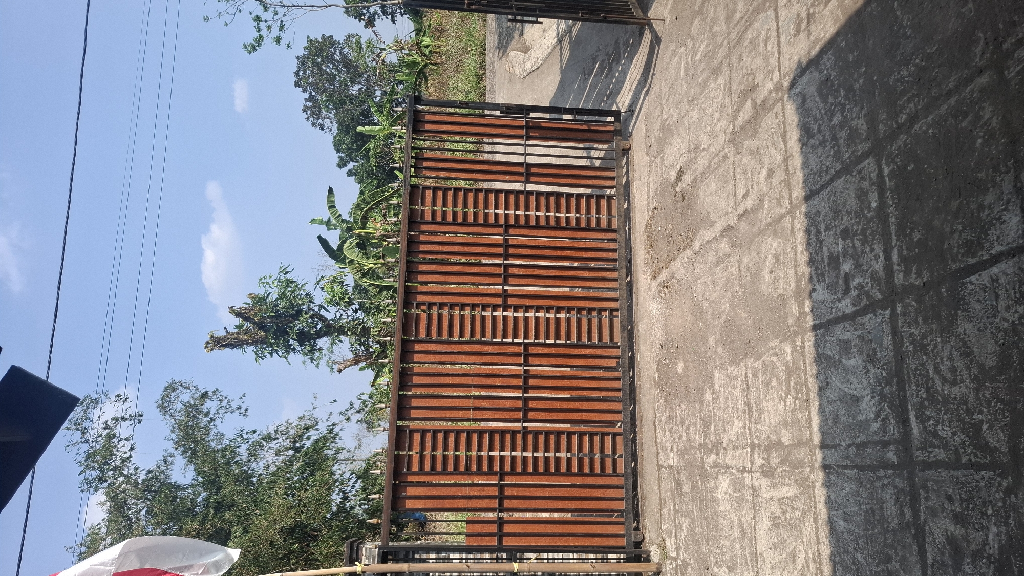 A medium shot captures a closed, wooden and metal gate in Cibinong Hilir, Indonesia. The gate is made of vertical wooden planks with horizontal slats, set within a black metal frame. To the left of the gate, a flagpole flies a white and red flag. Behind the gate, various trees, including banana trees, are visible. The ground in front of the gate is paved with concrete, showing signs of wear and tear, with a shadow cast across it from an unseen object. The sky above is clear and blue with a few scattered clouds. A portion of another black metal gate is visible on the far right of the image.