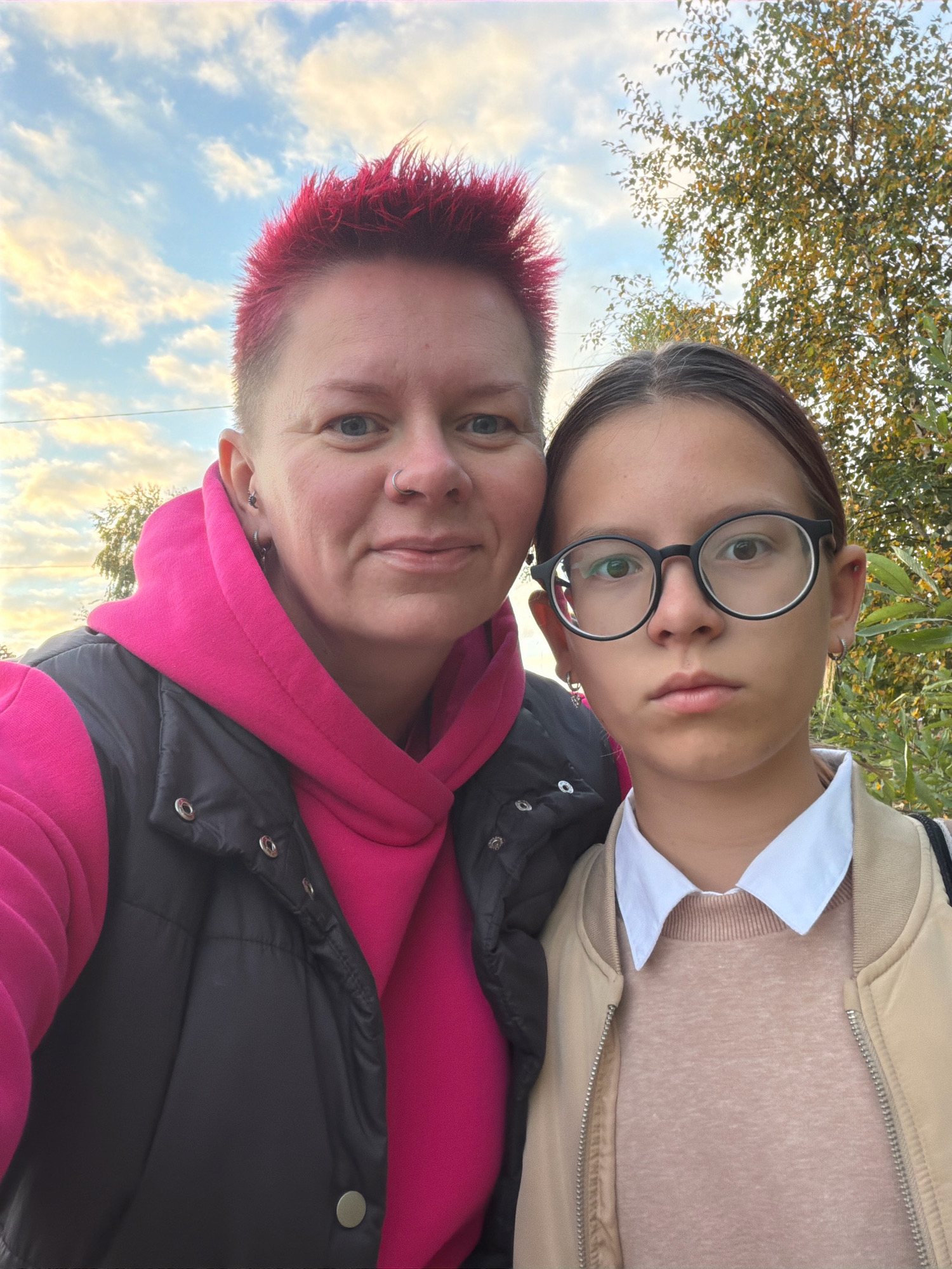 A close-up, waist-up selfie features two individuals, presumably mother and daughter, outdoors with a cloudy sky and autumn foliage in the background. The person on the left, identified as a woman, has bright pink, spiky hair, blue eyes, a nose ring, and is wearing a bright pink hoodie under a dark grey vest. The person on the right, identified as a girl, has brown hair pulled back, is wearing round black-rimmed glasses, a white collared shirt, and a beige bomber jacket over a light brown sweater. The lighting suggests it is daytime, possibly late afternoon. The backdrop includes trees with yellowing leaves, indicative of an autumn setting. The exact location within Chaikovsky, Russia, cannot be determined from the visual information provided.