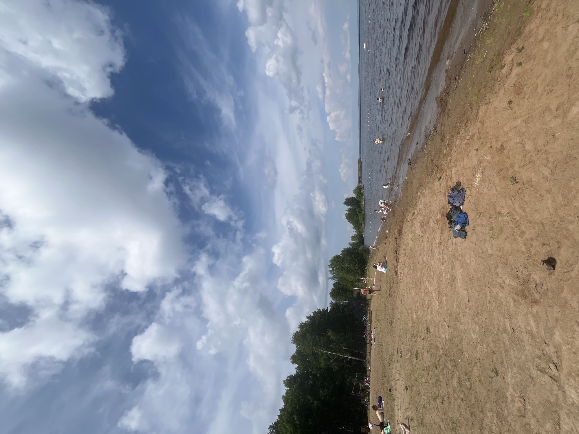 An outdoor, eye-level, full shot captures a sandy beach with a body of water on a cloudy, bright day. The sky is filled with large, fluffy white clouds against a backdrop of deep blue. The foreground is dominated by tan sand, with scattered green weeds. In the mid-ground, to the left, is a line of dense green trees. Further into the scene, several people are visible on the sand and in the water. Some individuals are lying on towels or sitting. A few people are wading in the water, and others are swimming. There are several bicycles parked near the trees. In the water, a small white boat is visible in the distance. To the far left, a volleyball net can be seen. Near the center of the foreground, a pile of blue and black bags lies on the sand.