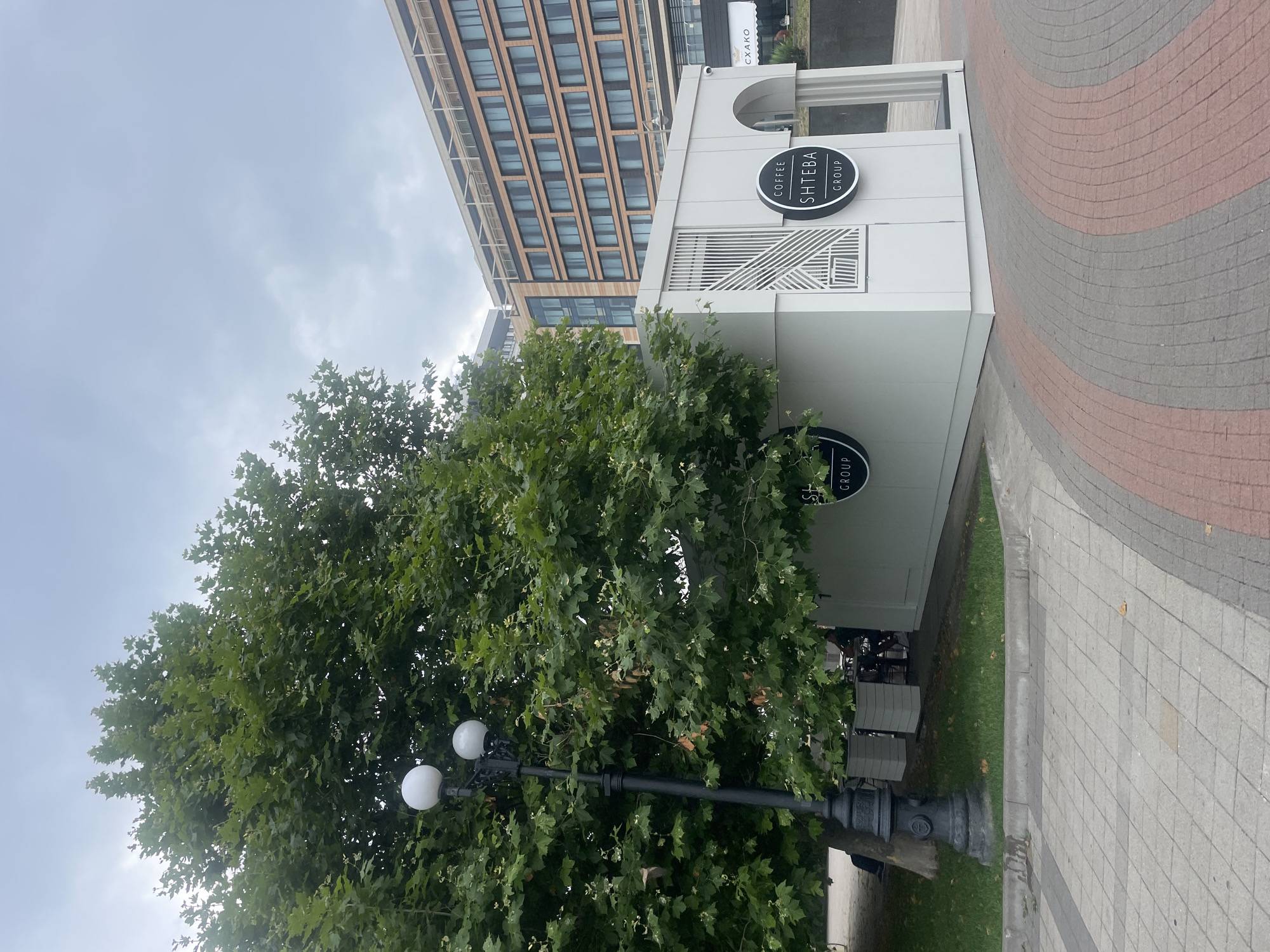 An exterior shot captures a modern, white coffee kiosk situated on a paved walkway in Novorossiysk, Russia. A large tree with lush green foliage is positioned to the left of the kiosk, partially obscuring it. A vintage-style lamppost with two globes stands near the tree. The kiosk features a minimalist design with geometric decorative elements on one side and an arched entrance on another. Two circular logos with the text "COFFEE SHTEBA GROUP" are visible on the kiosk. In the background, a multi-story building with a contemporary facade dominates the skyline under a cloudy sky. The walkway is paved with a patterned arrangement of grey and red bricks. A few people are seated at tables outside the kiosk, and one person is partially visible sitting on a bench near the tree.