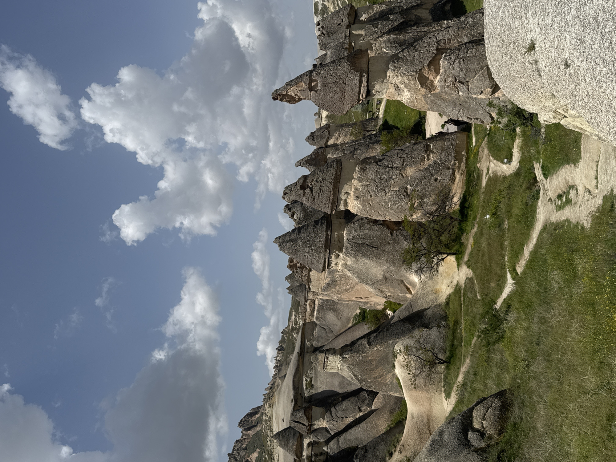 The image shows a landscape in Cappadocia, near Avanos, Turkey. The scene consists of fairy chimney rock formations, interspersed with patches of green vegetation and dirt paths. The sky is partially covered with white and grey clouds. There are no visible people. The image is taken from an elevated perspective.