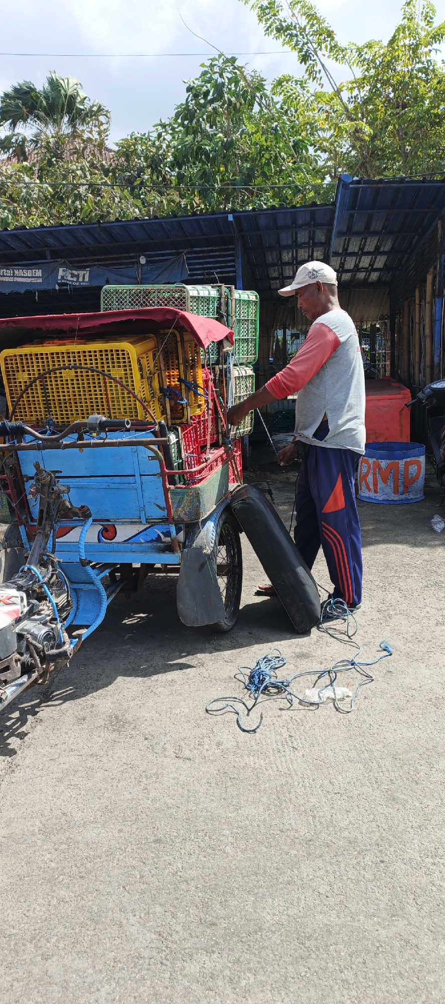 An outdoor scene in Puger, Indonesia, features a man in a white cap and a grey and pink long-sleeved shirt with blue pants with red stripes standing next to a three-wheeled vehicle. The vehicle is blue and red, laden with yellow and red crates. A black tire is leaning against the vehicle's rear. Blue rope lies coiled on the ground in front of the vehicle. In the background, a dark roof structure extends over a collection of green and white crates. Trees with lush foliage are visible above the roof. A blue cylindrical container with "RMP" spray-painted on it is situated to the right of the vehicle. The ground is paved with rough concrete.