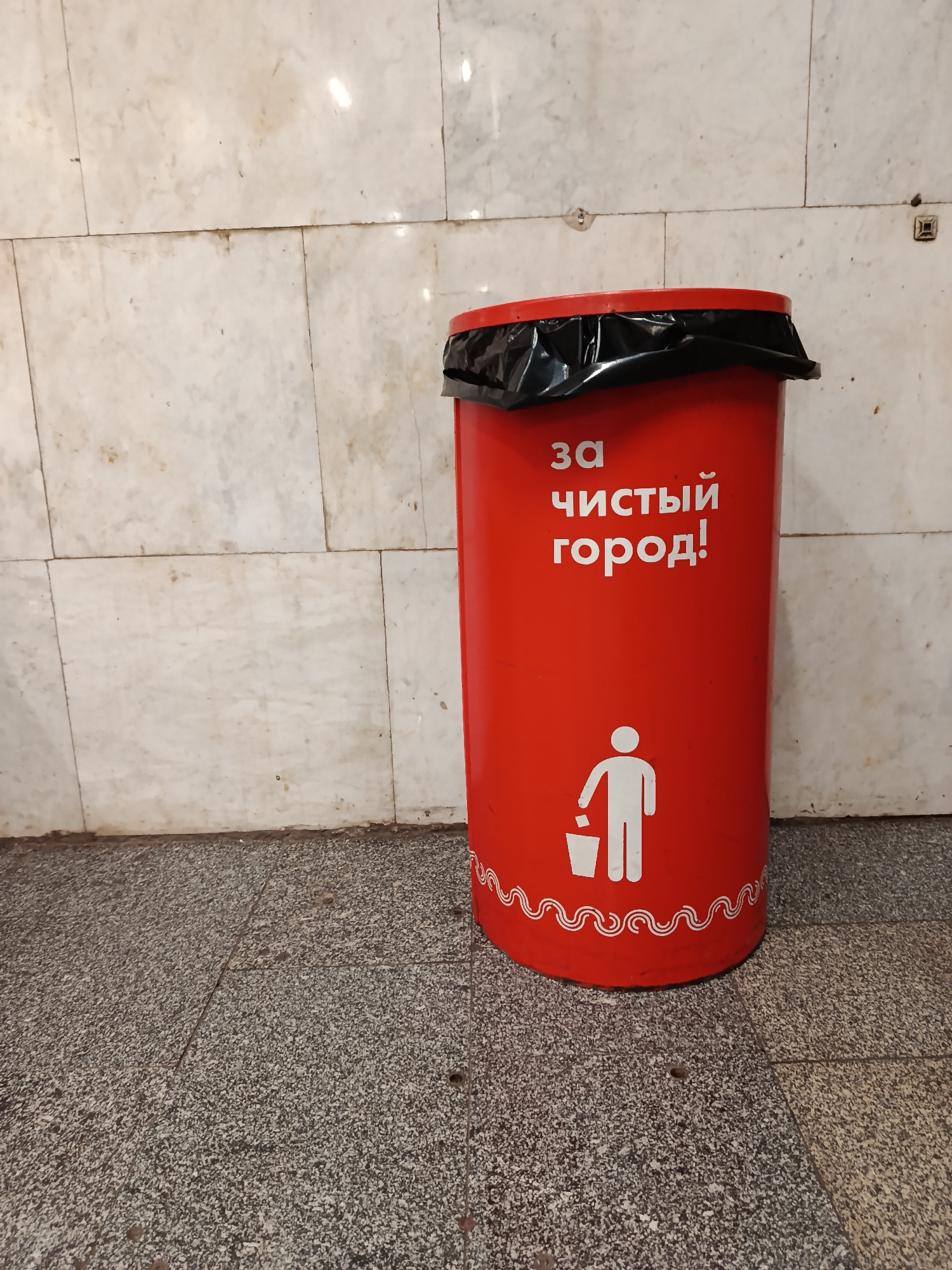 A red, cylindrical trash can stands against a marble-tiled wall in what appears to be a public space, possibly in the city of Moscow, Russia. The trash can is lined with a black plastic bag. White lettering in Cyrillic script reads "за чистый город!" (for a clean city!). Below this, a white pictogram shows a figure dropping trash into a receptacle. A wavy, decorative line encircles the bottom of the can. The floor is composed of gray granite tiles.