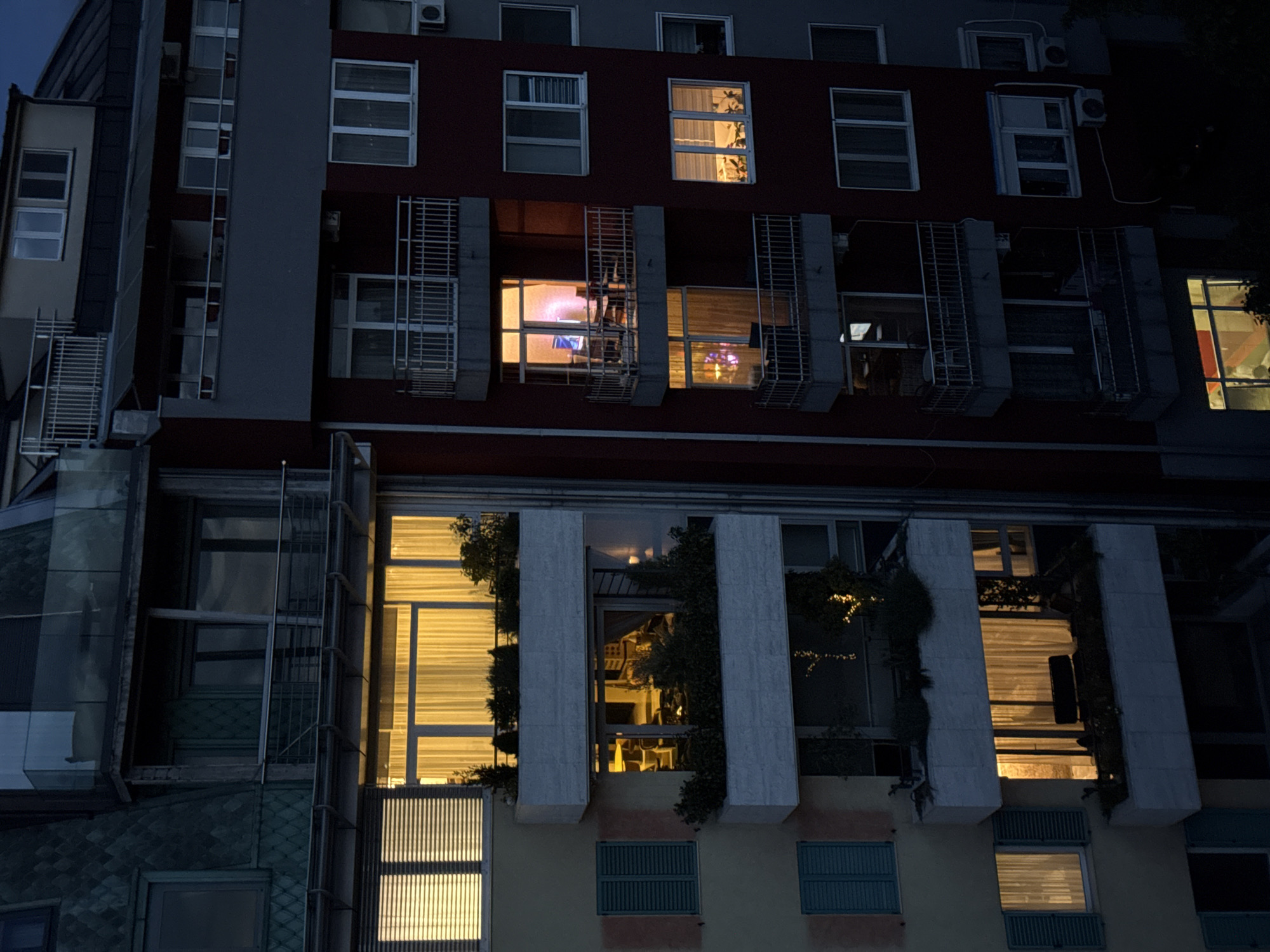 The image depicts a multi-story building at night in Vracar Urban Municipality, Serbia. The building has a mix of architectural styles, with glass panels on the lower floors and a more traditional facade on the upper floors. Some windows are illuminated, suggesting occupancy. Shutters and metal grates are visible on some windows, adding to the complexity of the building's design. Climbing plants are growing on parts of the facade. The lighting suggests it is dusk or night, and the overall scene is quiet and urban. There are no people visible in the image.