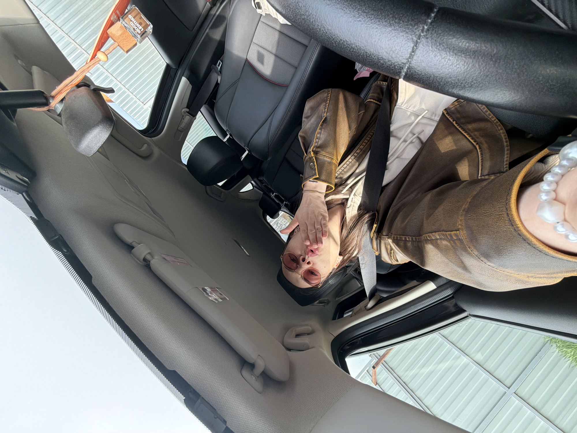 A person is in the driver's seat of a car. The person is wearing sunglasses, a brown jacket, and a pearl necklace. The person is blowing a kiss towards the camera. The car is parked in Chaikovsky, Russia. The interior of the car is visible, including the dashboard, seats, and rearview mirror. There is a car air freshener hanging from the rearview mirror. The sun visor is down on the driver's side. The environment outside the car includes other buildings and possibly the sky.