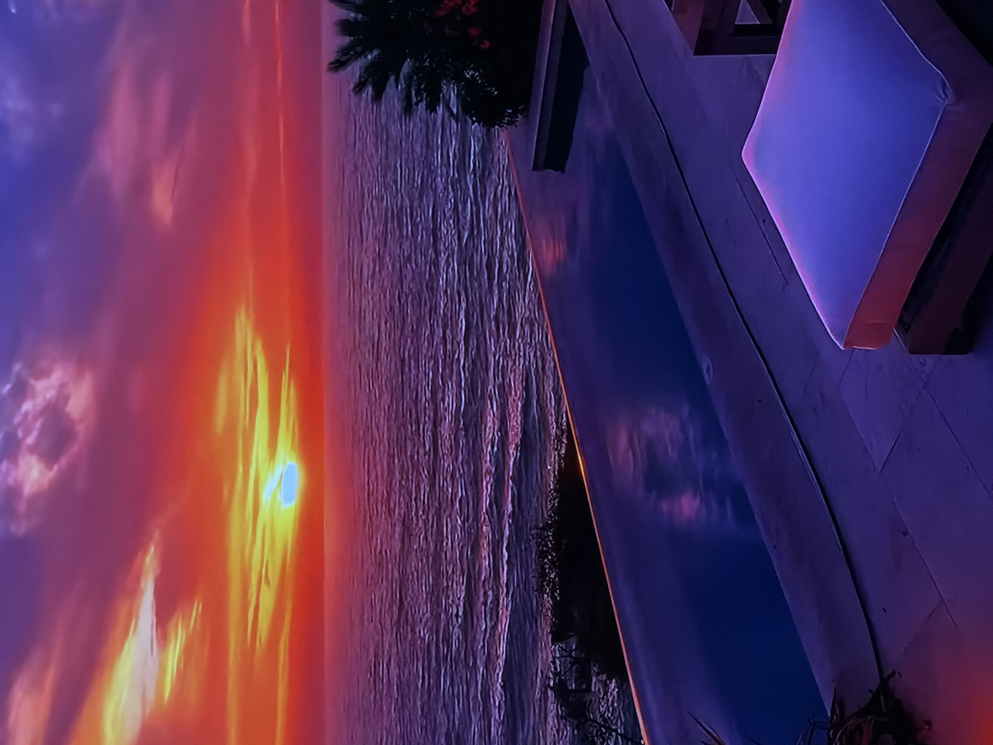 The image captures a vibrant and dramatic sunset scene from an elevated perspective, likely from a resort or villa overlooking a large body of water. The overall impression is one of serene beauty and luxury.
In the foreground, a tranquil swimming pool runs parallel to the horizon. Its calm surface reflects the deep blue and purple hues of the twilight sky. Adjacent to the pool, a section of an outdoor deck is visible, featuring dark-framed lounge furniture with a prominent, bright white or light blue cushion that stands out against the darker surroundings, suggesting comfort and relaxation. Dark, silhouetted foliage, possibly palm fronds, frames the upper right portion of the foreground, adding to a tropical ambiance.
The mid-ground is dominated by the vast expanse of a sea or ocean. Its surface is textured with small ripples, reflecting the muted purples and blues of the sky. A dark, indistinct coastline or landmass with more scattered foliage can be seen where the water meets the sky on the right side of the frame.
The background is a breathtaking display of a sunset. A brilliant, intensely glowing sun, appearing almost white or light blue at its core, is visible low on the horizon, partially obscured by illuminated clouds. The sky around the sun erupts in a fiery palette of vivid oranges, yellows, and reds, which gradually transition upwards into rich purples and deep blues towards the upper left. Scattered clouds are visible, catching the last light of the day and enhancing the atmospheric effect.
The time of day is clearly sunset or twilight, characterized by the strong contrasting colors and dramatic natural lighting. No people or specific activities are depicted. There is no visible text. The tropical foliage, large body of water, and luxurious poolside setting strongly suggest a coastal, warm-climate location.